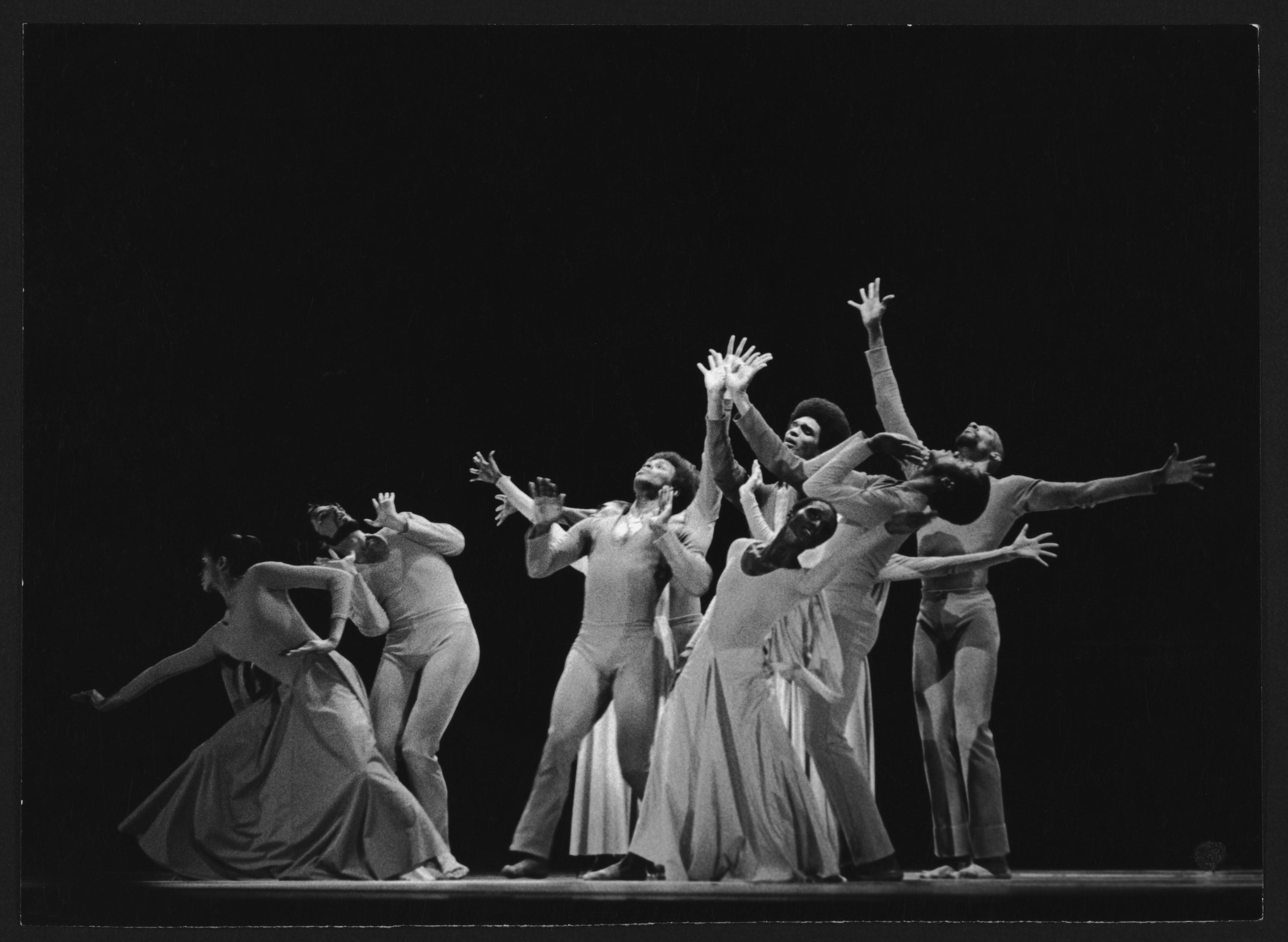 A black and white photo of a performance featuring Donna Wood, Peter Woodin, Freddy Romero, Sylvia Waters, Clive Thompson, Melvin Jones, and John Parks in Choral Dances. The dancers are grouped closely together, striking expressive and dynamic poses with their arms extended and faces turned upward. They are wearing light-colored costumes, and the background is dark, highlighting the dancers.