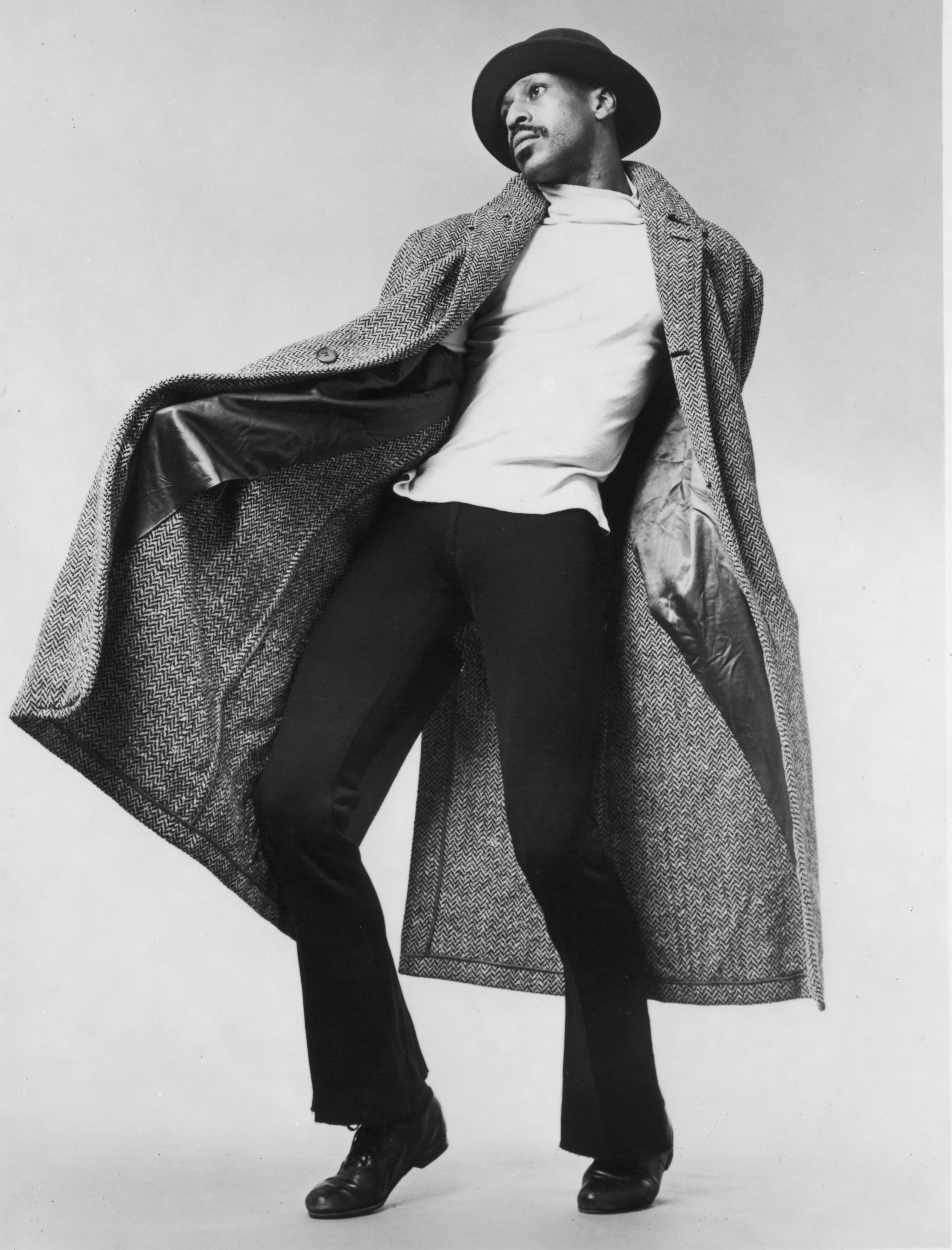 A black and white photo of dancer Dudley Williams in mid-performance. He is wearing a large, herringbone-patterned coat, a light shirt, dark pants, and a hat. He is captured in a dynamic pose, with the coat flaring open and a thoughtful gaze to the side.