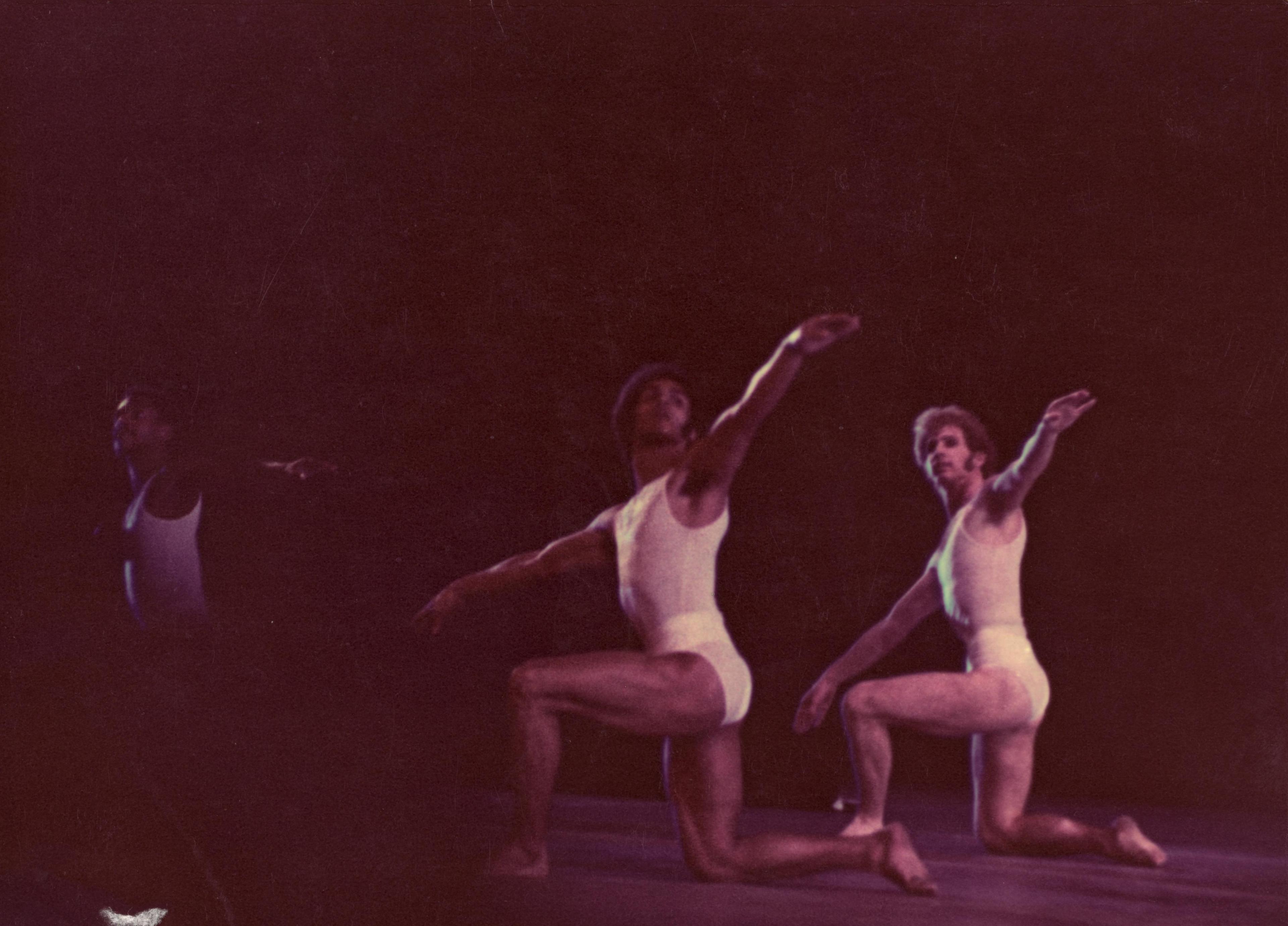 A color photo of dancers Dudley Williams, Hector Mercado, and Leland Schwantes in mid-performance. They are kneeling on one knee with arms extended, wearing white costumes. The lighting is dim, creating a reddish hue over the image.