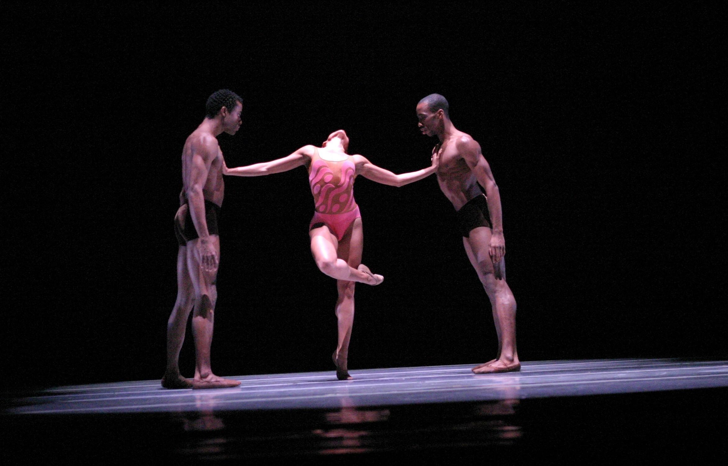 A color photo of three dancers in mid-performance. The central dancer, wearing a pink leotard, is balanced on one leg, leaning back with arms extended, supported by two male dancers in dark shorts. The male dancers stand on either side, holding her arms. The background is dark, emphasizing the dancers. 