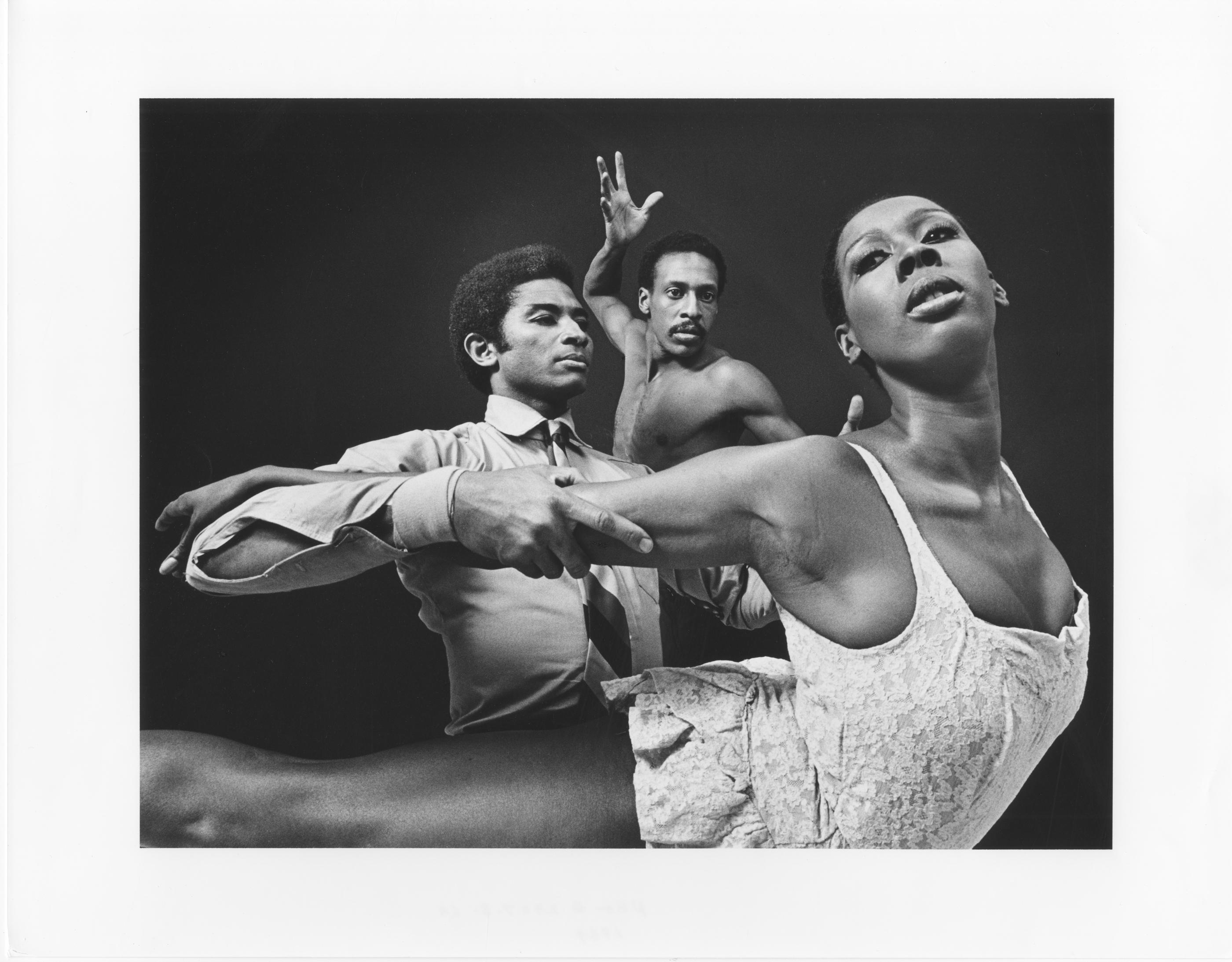 A black and white photo of dancers Kelvin Rotardier, Dudley Williams, and Judith Jamison in mid-performance. Judith Jamison is in the foreground, extending her body forward in a dynamic pose, wearing a lacy dress. Kelvin Rotardier, dressed in a shirt and tie, is holding her arm, while Dudley Williams, shirtless, is behind them with an arm raised. The background is dark, highlighting the dancers.