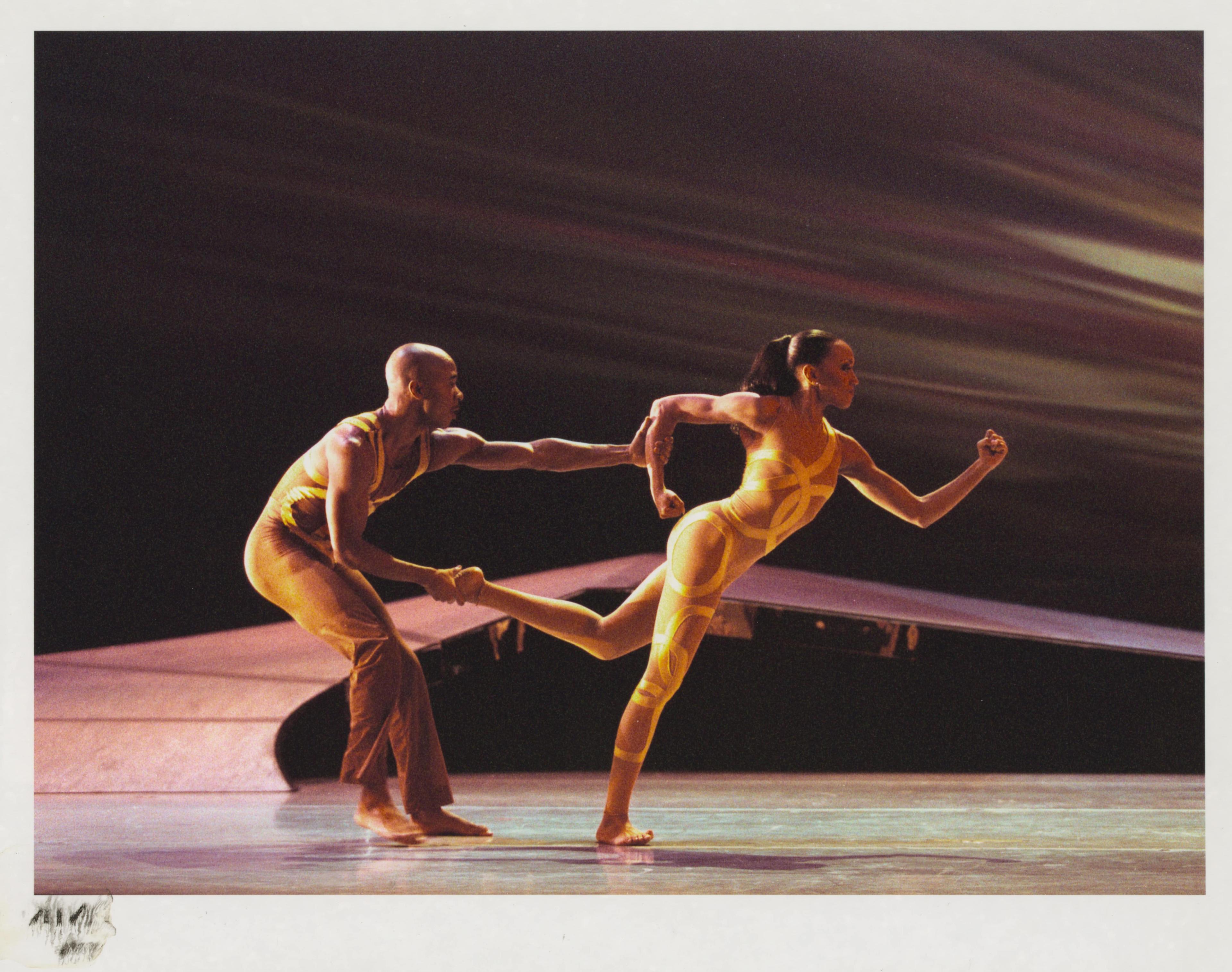 Color photo of Matthew Rushing and Linda-Denise Fisher-Harrell performing in HERE...NOW. Matthew is wearing brown pants and a sleeveless top, holding Linda-Denise, who is in a yellow bodysuit with crisscross patterns, as she leans forward in a dynamic pose.