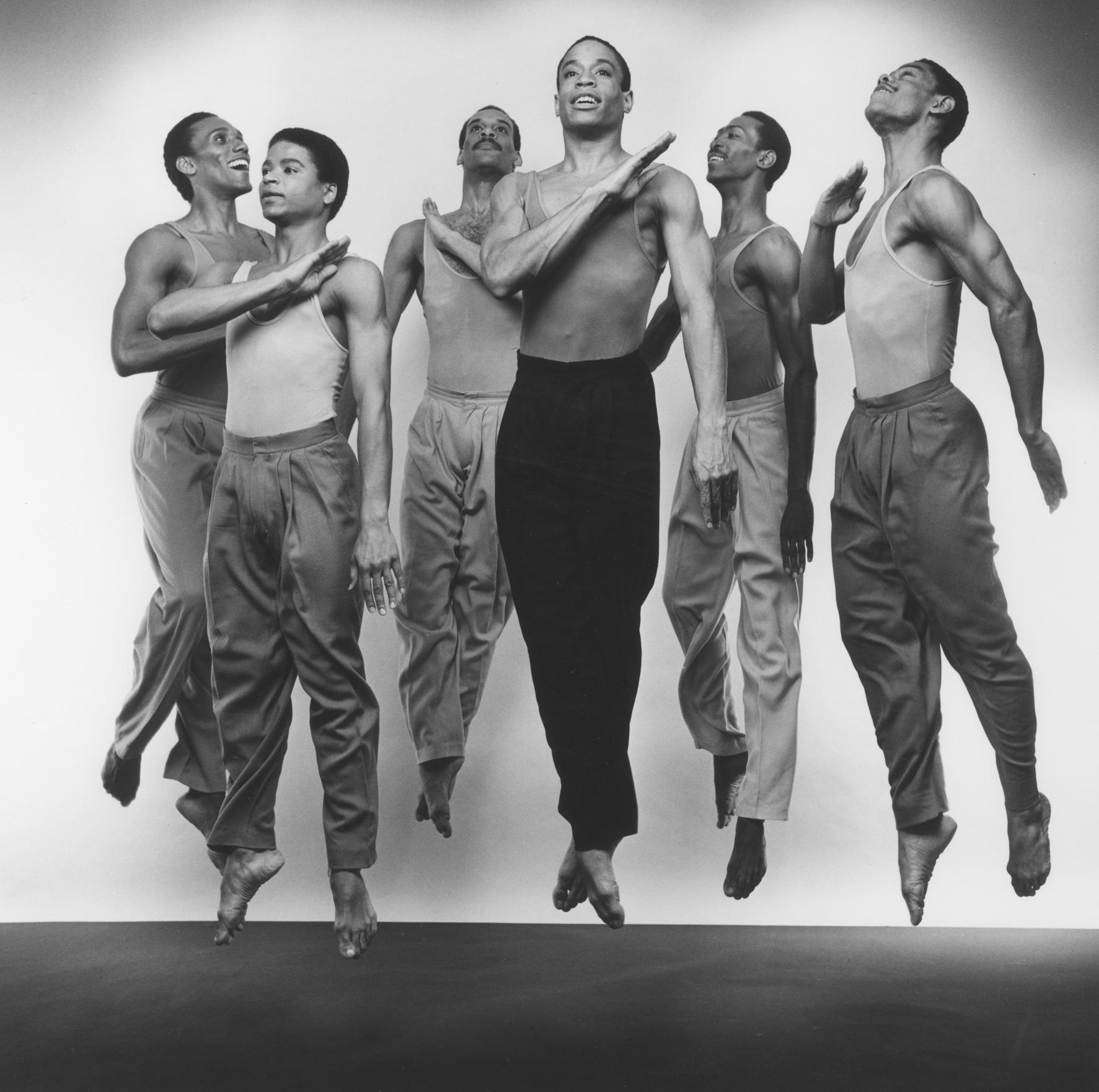 Black and white photo of six members of the company performing in Fever Swamp. The dancers are captured mid-jump, wearing sleeveless tops and trousers. The central dancer is shirtless with dark pants.