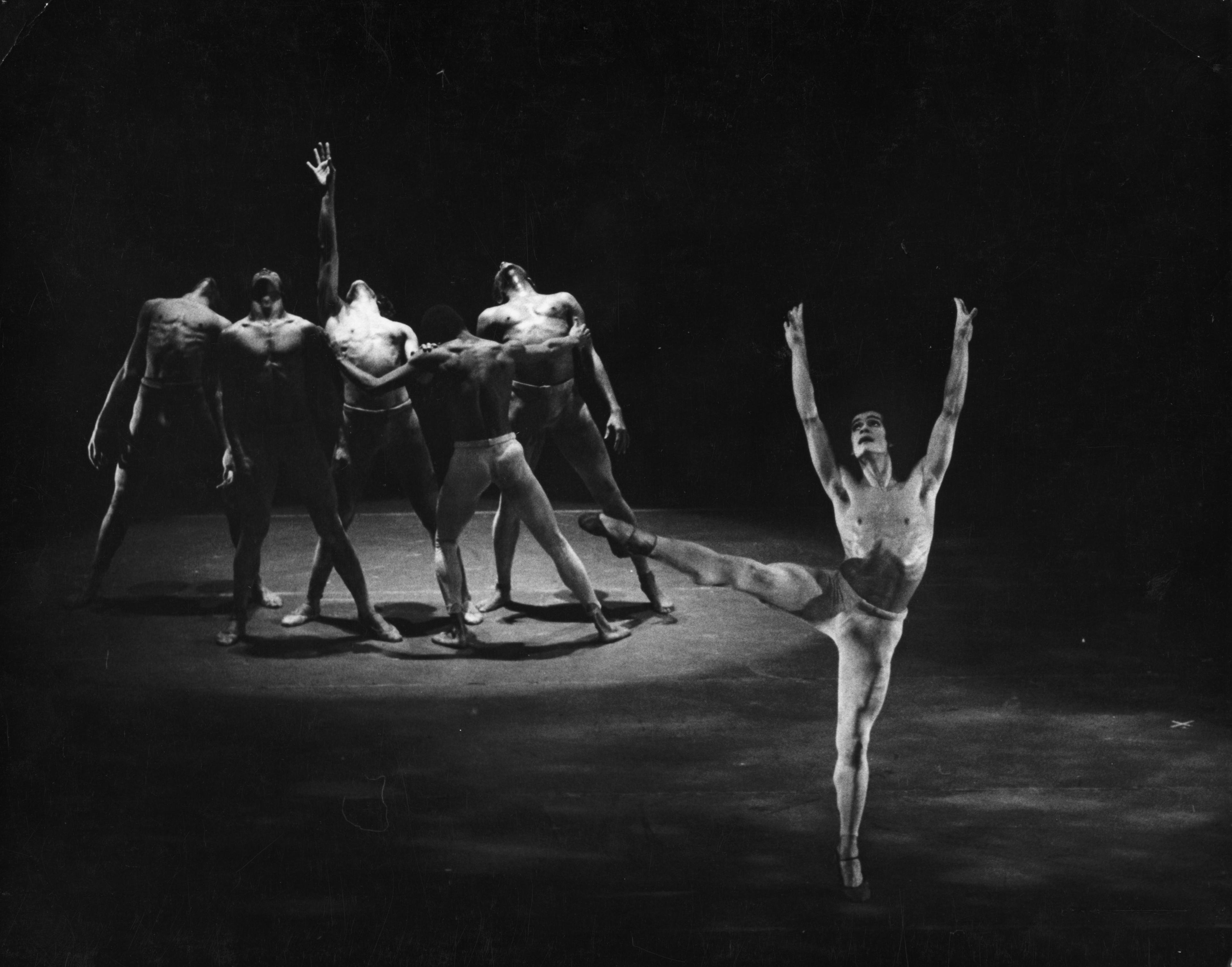 Black and white photo of Ramon Segarra performing in Archipelago. Ramon is in the foreground, balancing on one leg with arms raised and wearing minimal costume. In the background, a group of male dancers is huddled together in dramatic poses.