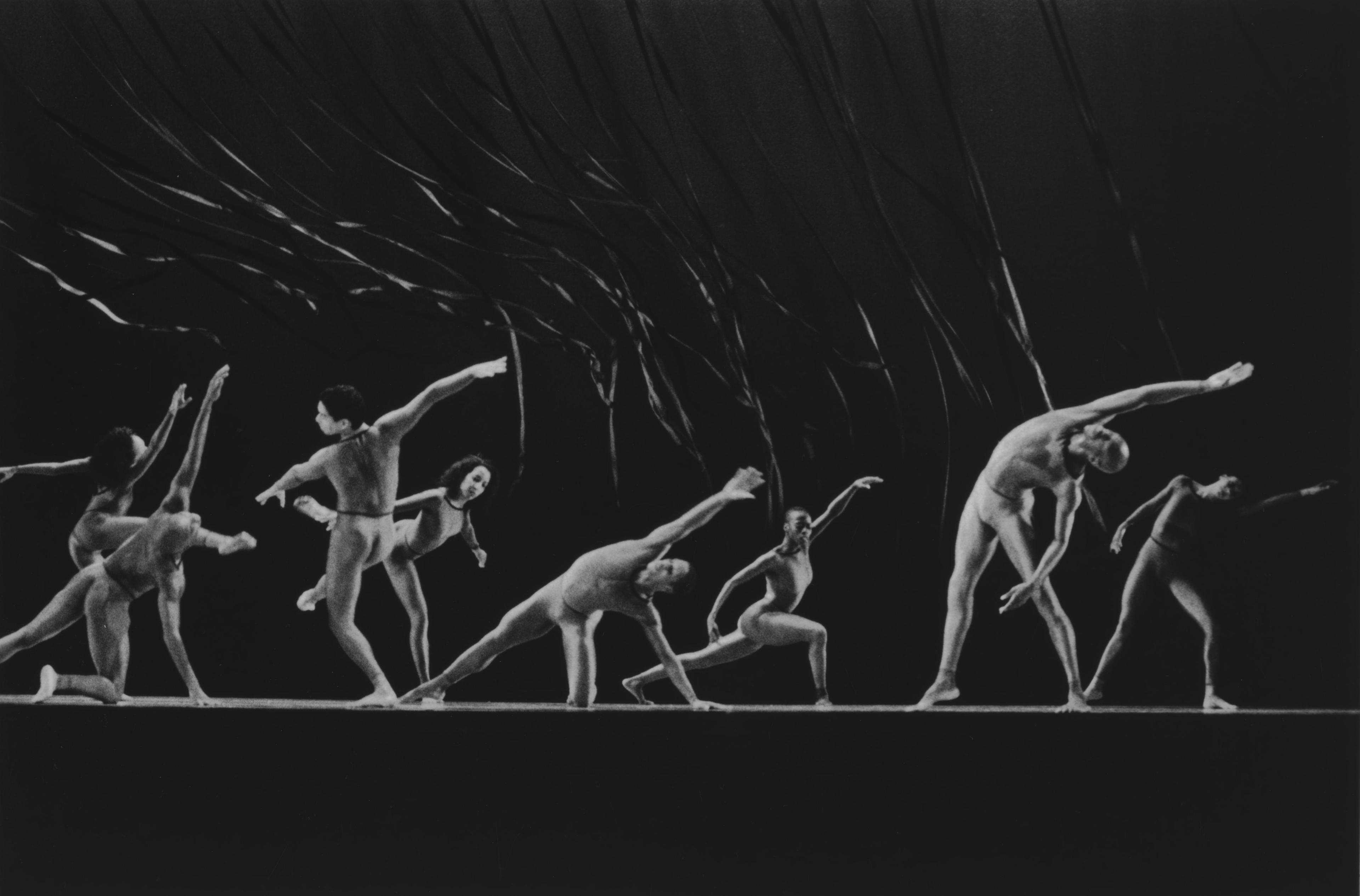 Black and white photo of the company performing in Cavalcade. The dancers are in various dynamic poses, stretching and leaning, with their arms extended. They wear minimal costumes. Ribbons or streamers are visible in the background, adding to the visual movement.