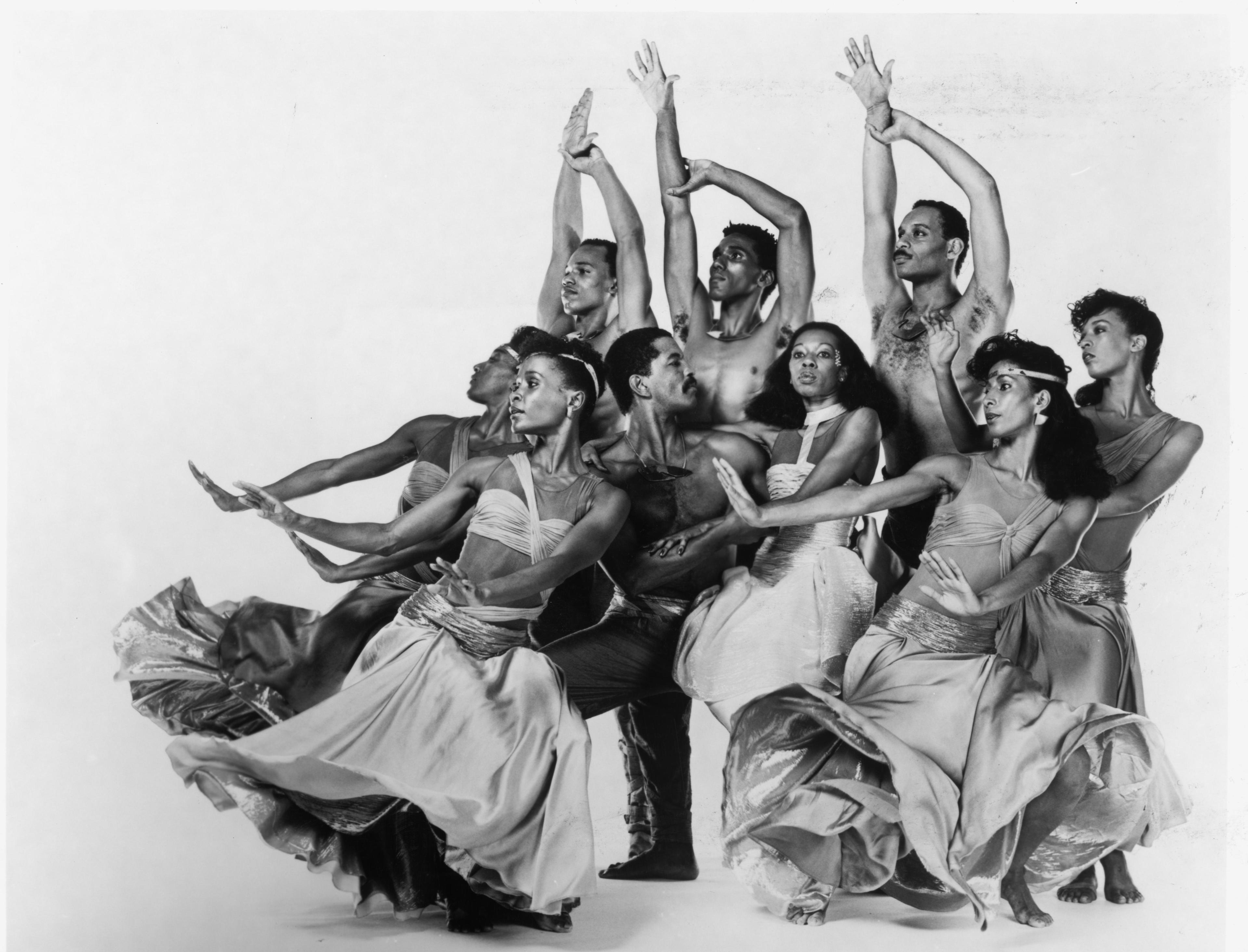 Black and white photo of the company performing in Caverna Magica. The dancers are arranged in a dynamic group pose, with flowing skirts and expressive arm movements. Some dancers are shirtless, while others wear fitted tops, capturing a sense of unity and energy.