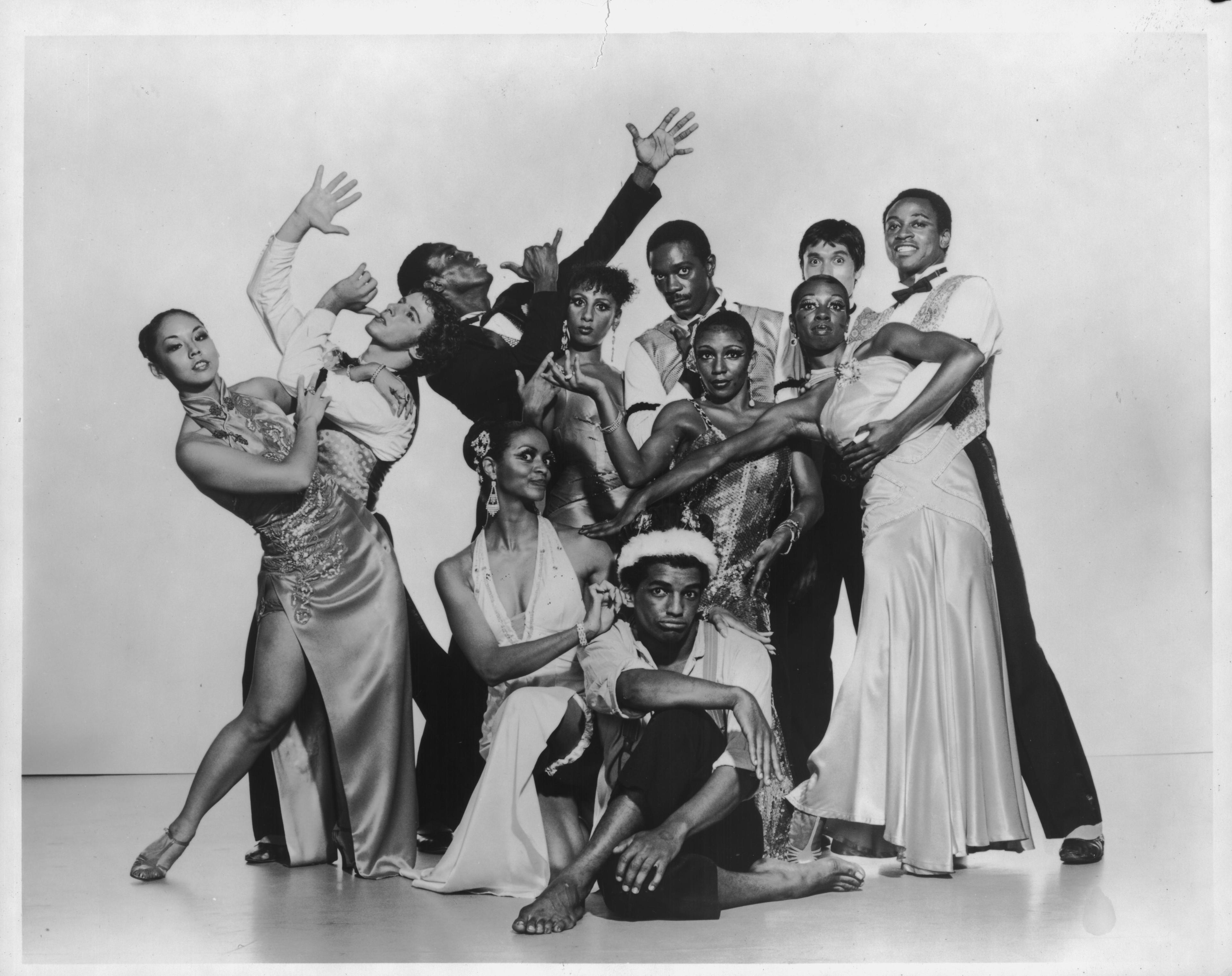 Black and white photo of the company performing in District Storyville. The dancers are dressed in elegant attire, with women in gowns and men in suits, striking expressive poses. The group is arranged in a lively formation, capturing a sense of drama and movement.