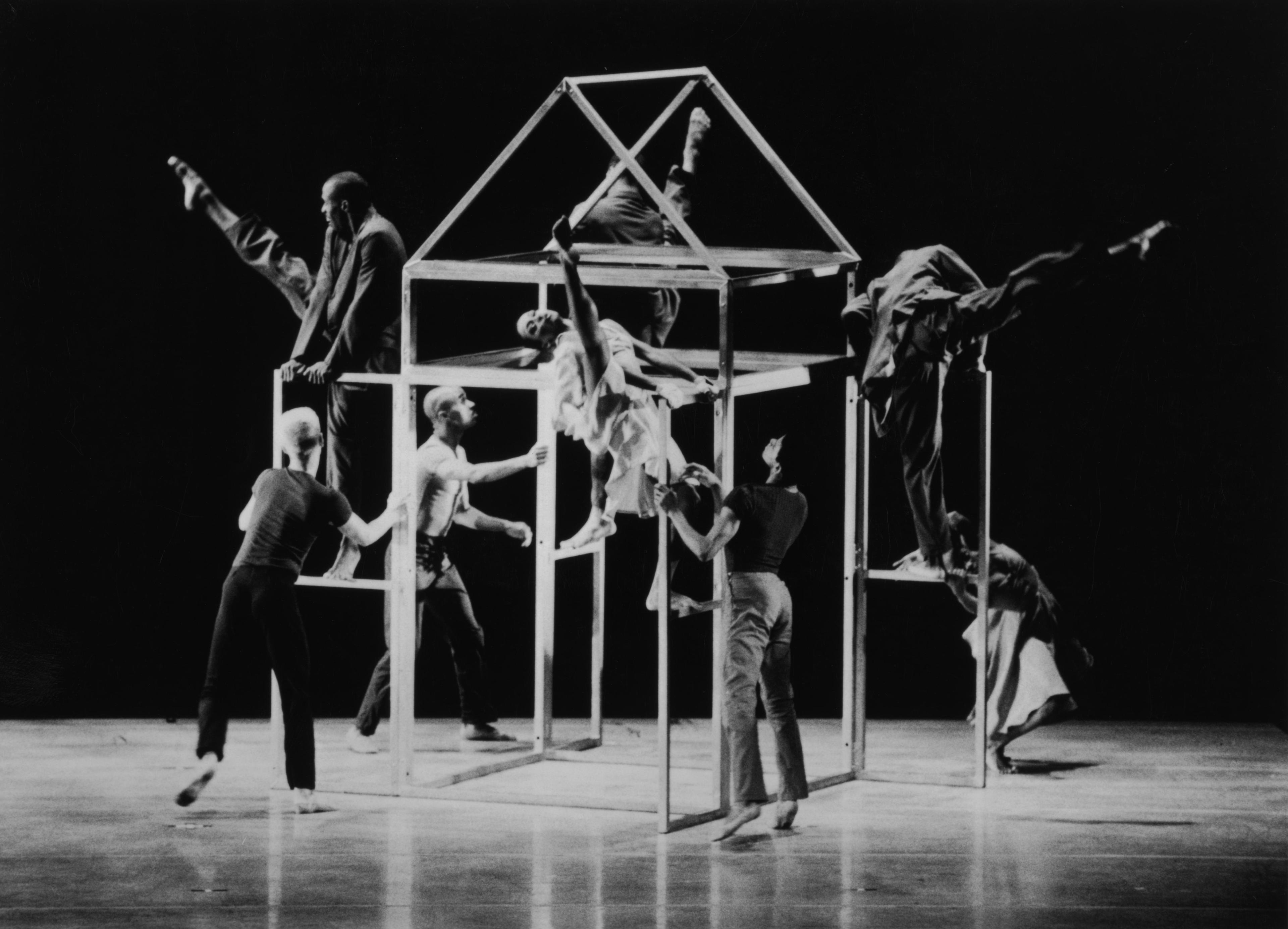 Black and white photo of the company performing in Fathers and Sons. The dancers are interacting with a frame structure resembling a house, with some climbing and others extending their limbs in dynamic poses. The scene captures a sense of movement and balance.