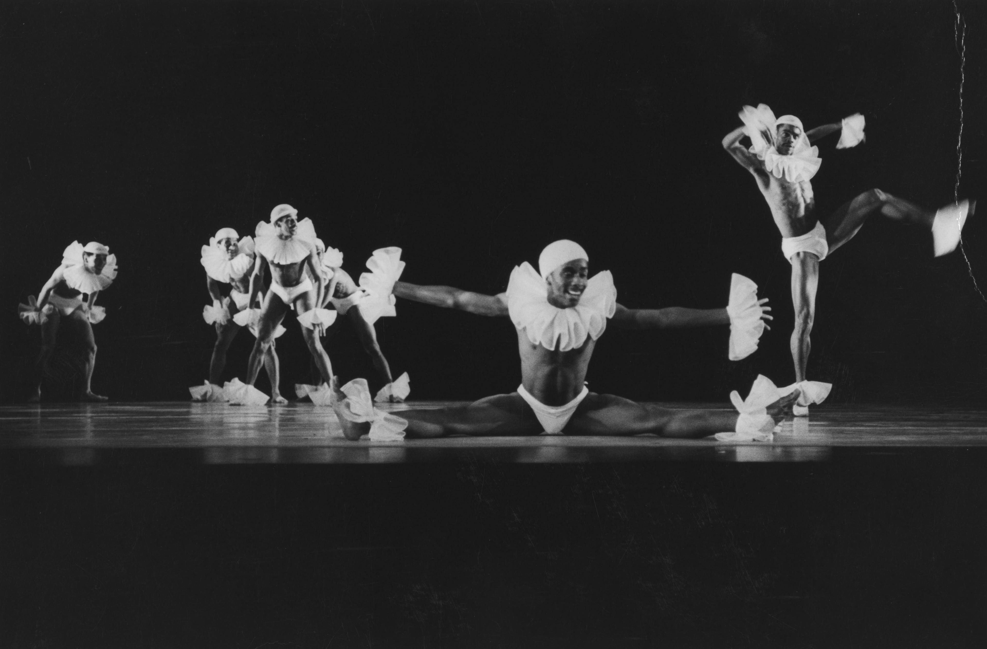 A black and white photograph of a dance performance featuring six dancers dressed in elaborate costumes with ruffled collars and cuffs. One dancer is performing a split on the floor, arms extended. Another dancer is captured mid-air with one leg raised. The other dancers are in various dynamic poses in the background.