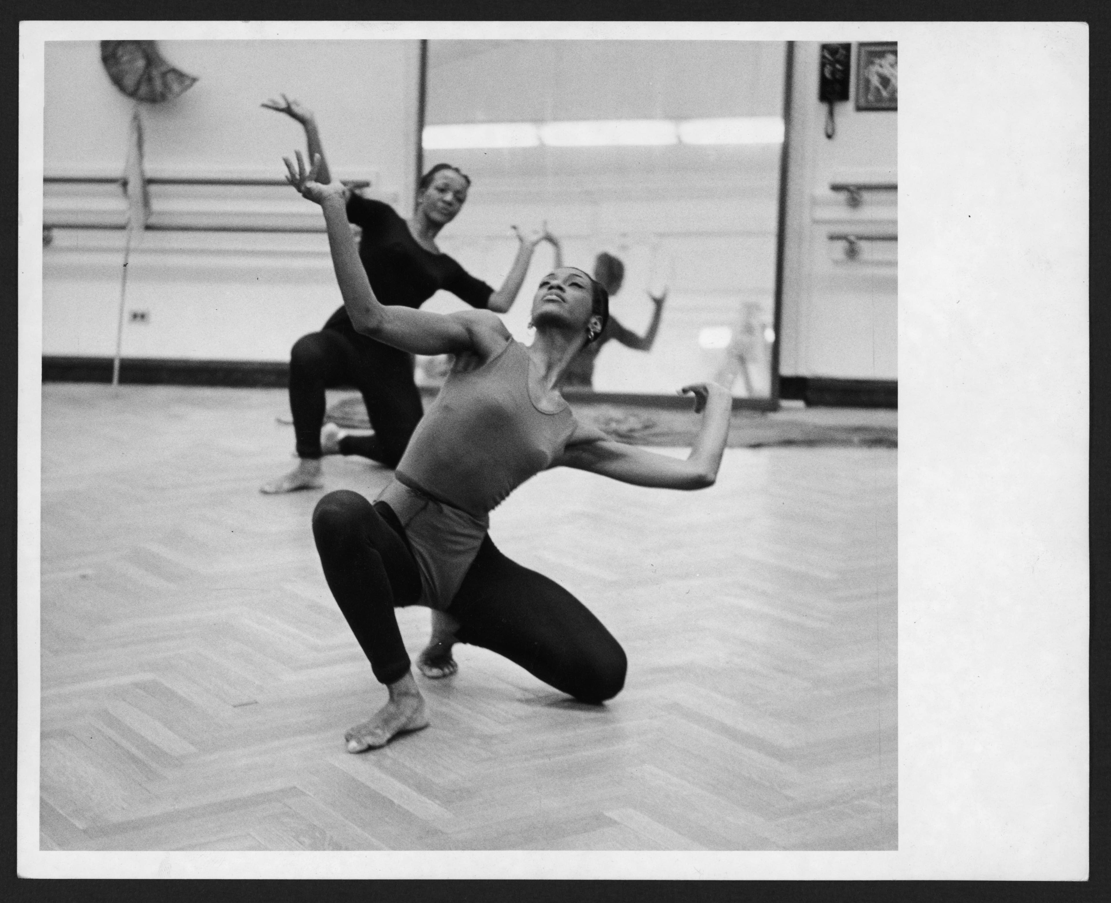 A black and white photograph of two dancers performing in a studio. The dancer in the foreground is kneeling on one knee with her arms raised in an expressive pose, looking upward. The dancer in the background is partially visible, also in a dynamic pose. The studio has a wooden floor and mirrors on the walls.