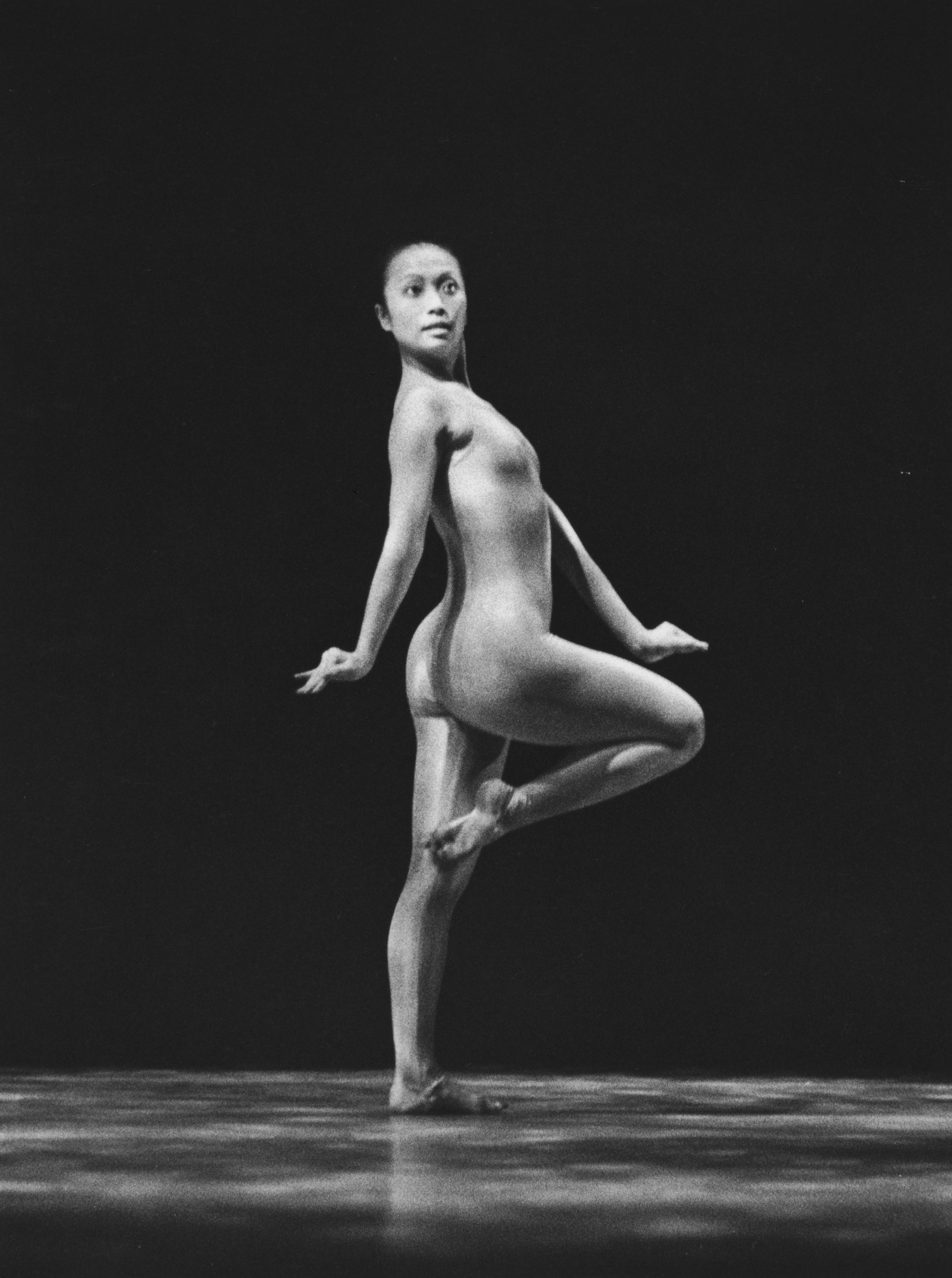 A black and white photograph of a dancer named Tina Yuan performing in a piece titled "Gazelle." She is captured in a graceful pose, standing on one leg with the other leg bent and her arms positioned elegantly. The background is completely dark, emphasizing her form.