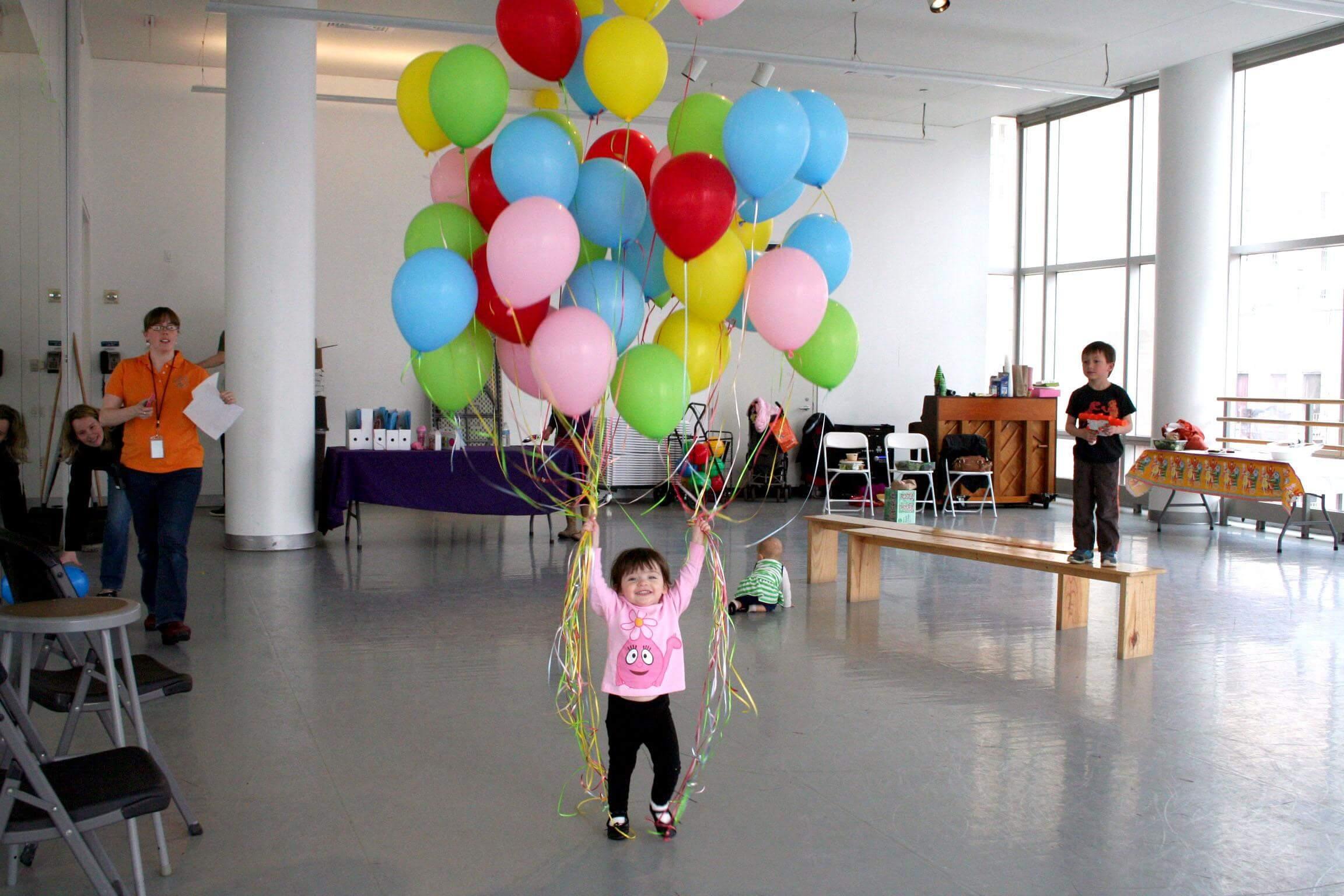 A little girl in a pink shirt and black pants holding a bouquet of blue, green, yellow, red, and pink balloons. She is in a dance studio with party decor in the background.