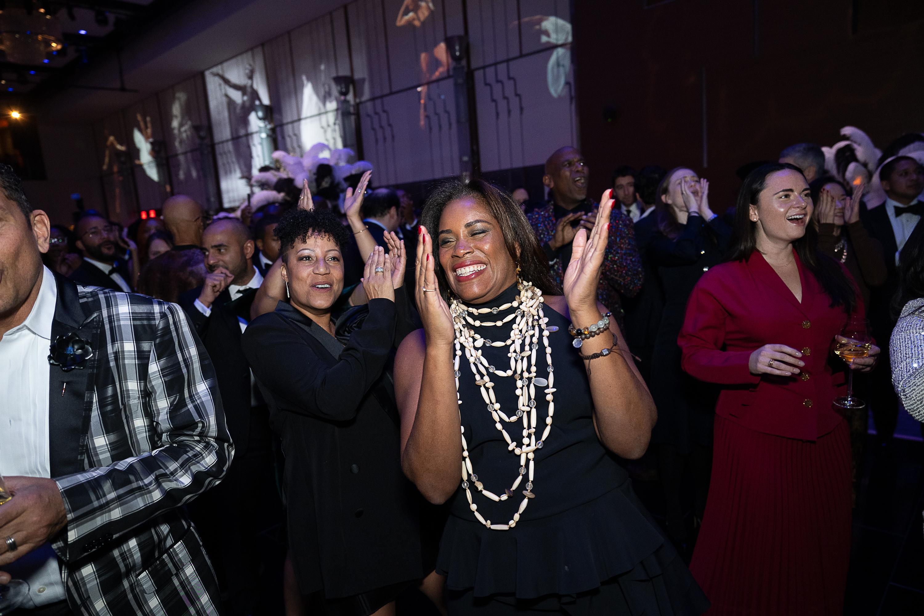 People dancing in a grand ballroom. There are two women in the middle of the image. They are clapping their hands and smiling. The woman slightly in the front of the image has a black dress on with many silver necklaces.