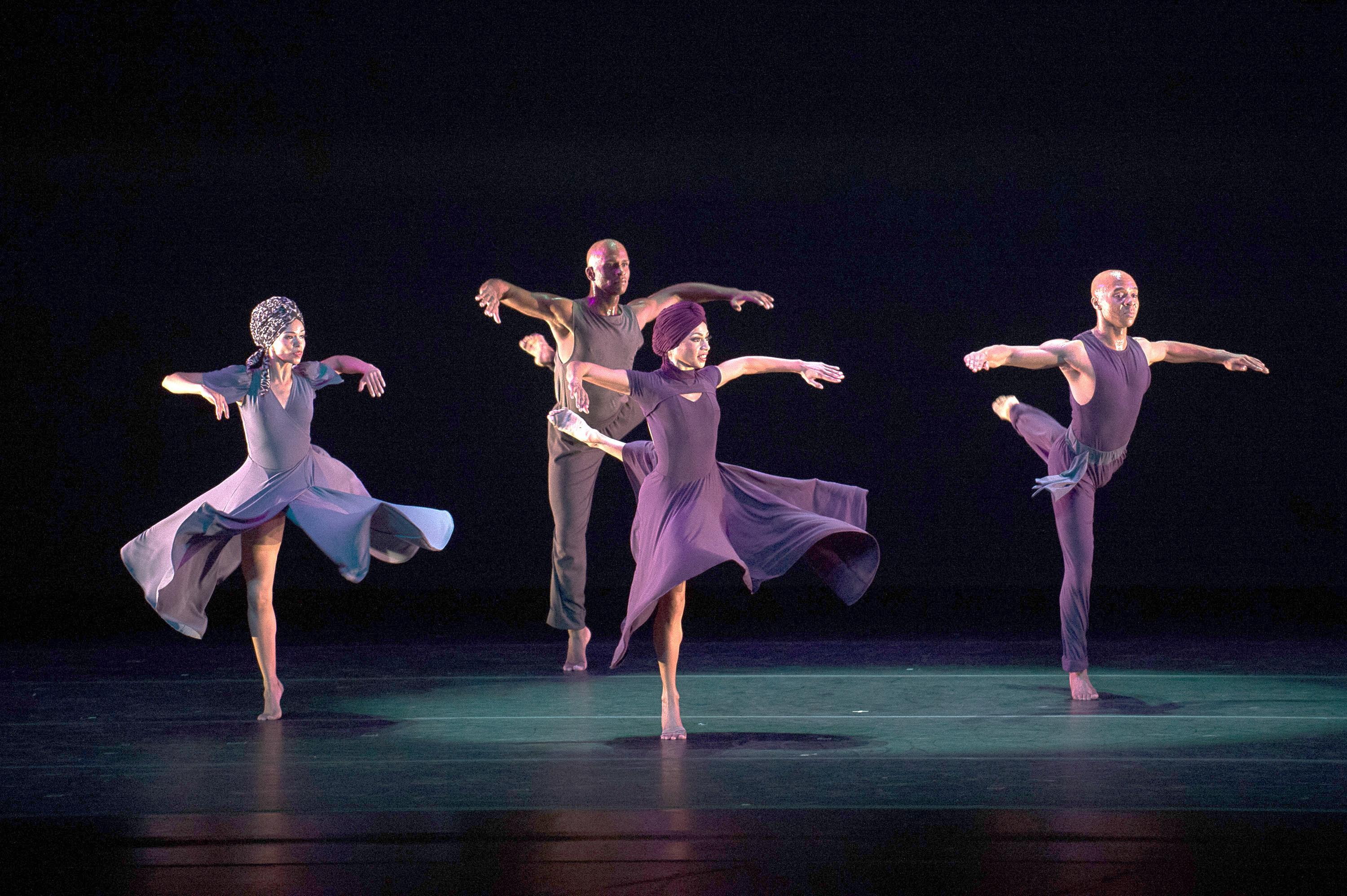 four dancers, two men, and two women stand in a square shape perched on one leg, arms out to the side.