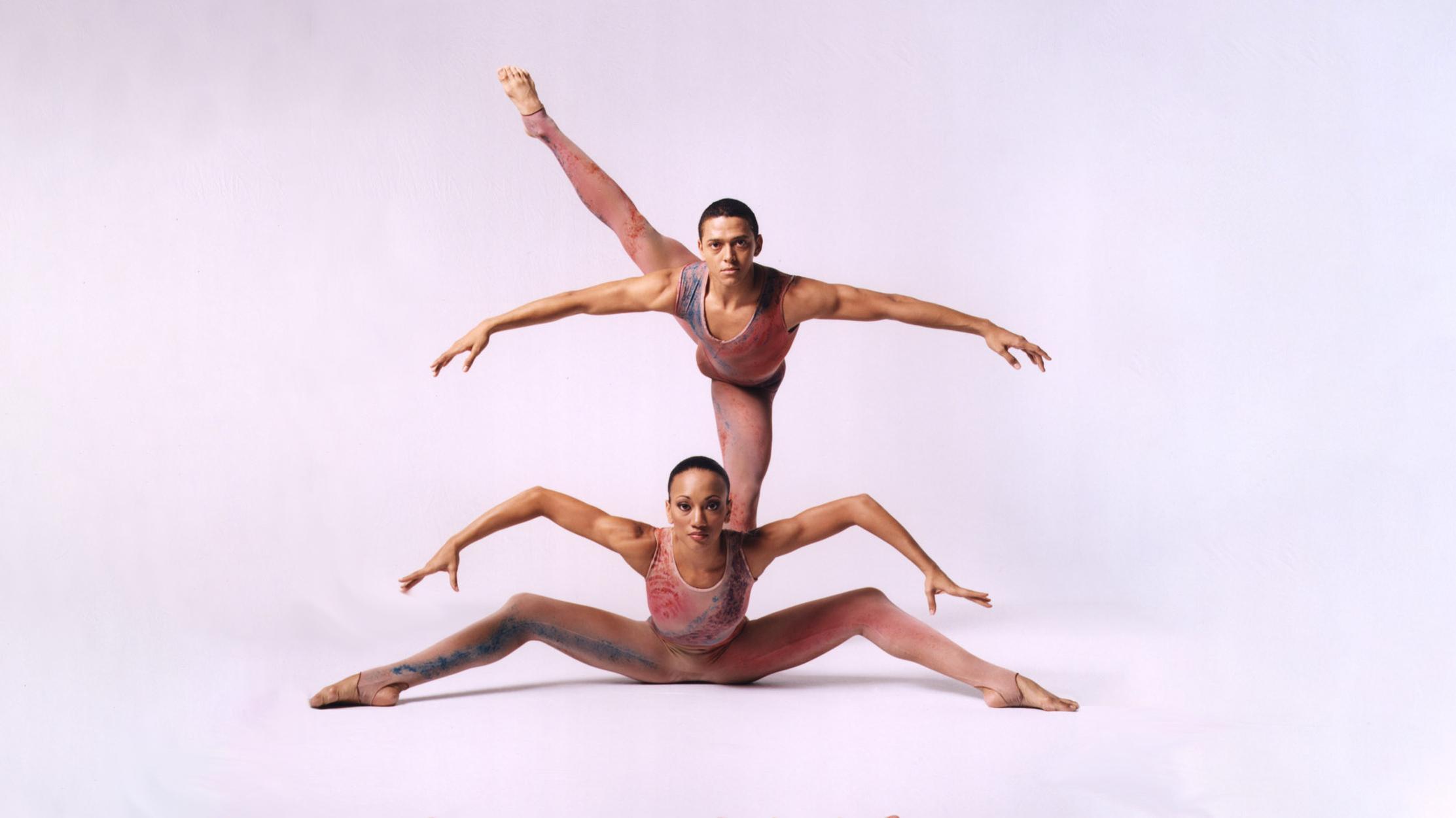 a woman sits on the ground with her legs and arms spread out and a man stands on one leg with othe other reaching out to the side, behind her on