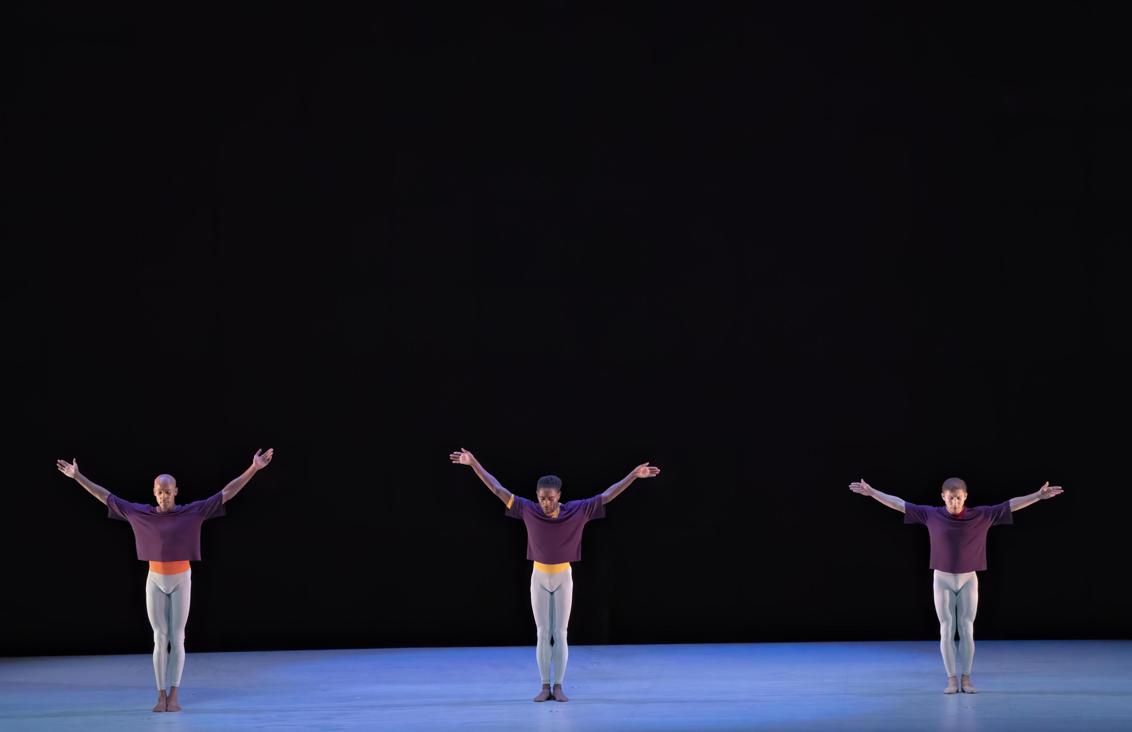 Three men wearing purple shirts and blue tights stand on a stage. next to each other with their arms reaching up above them.