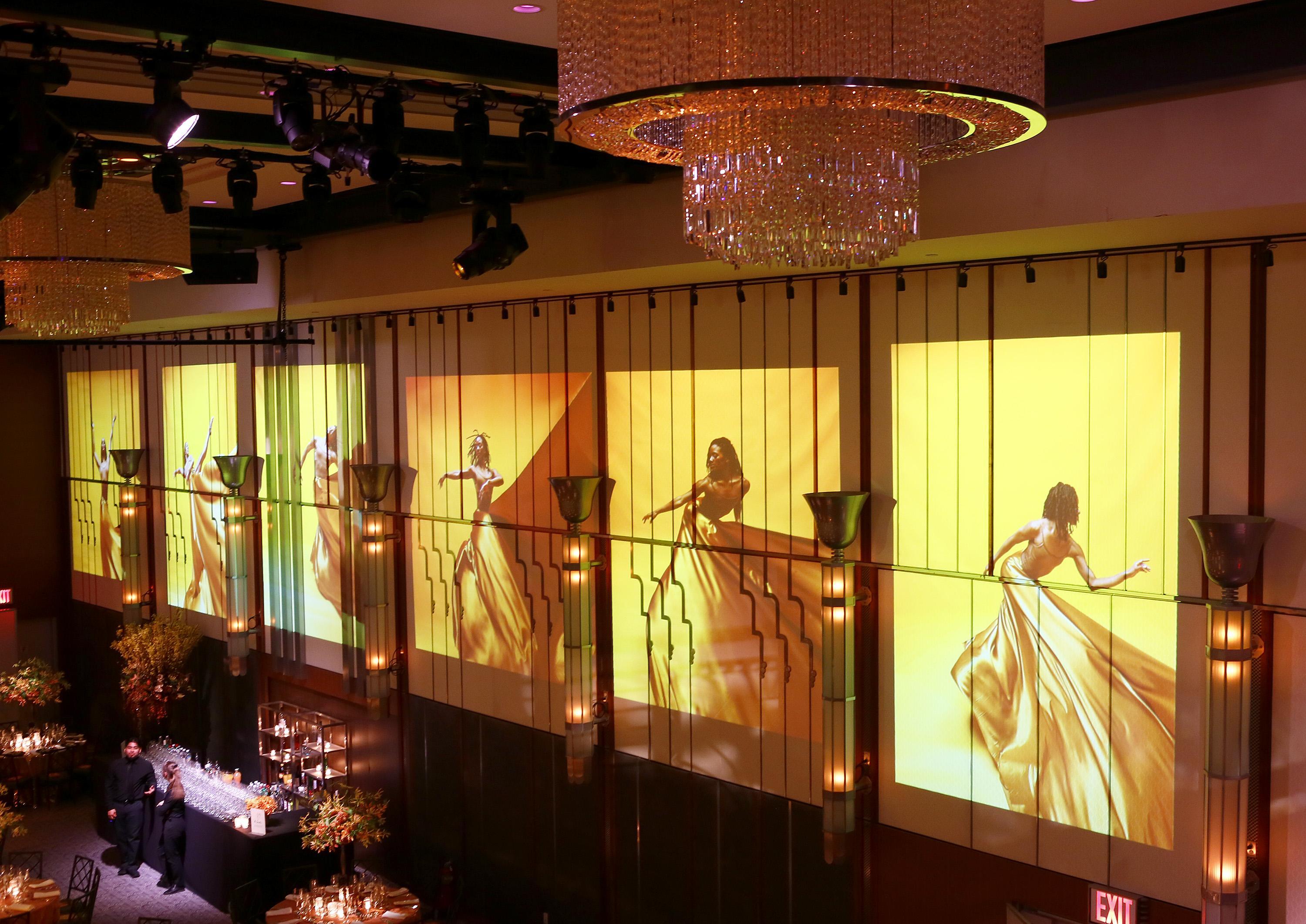 A ballroom with five different images of dancers projected onto the wall. There is a bar with glasses at the bottom of the image. There is a crystal chandelier at the top of the image.