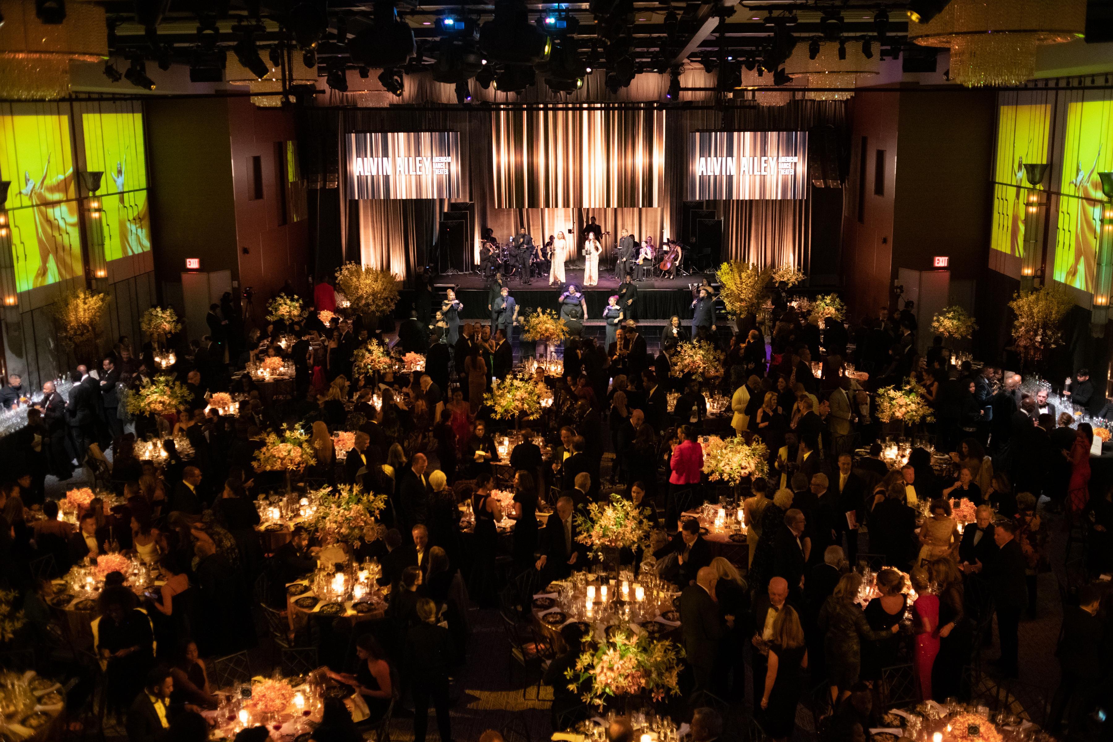 Ailey's 2022 Opening Night Gala. A wide shot of a grand ballroom. There are tables with arrangements and flowers, images of dancers projected on the walls, and a band warming up in the background.