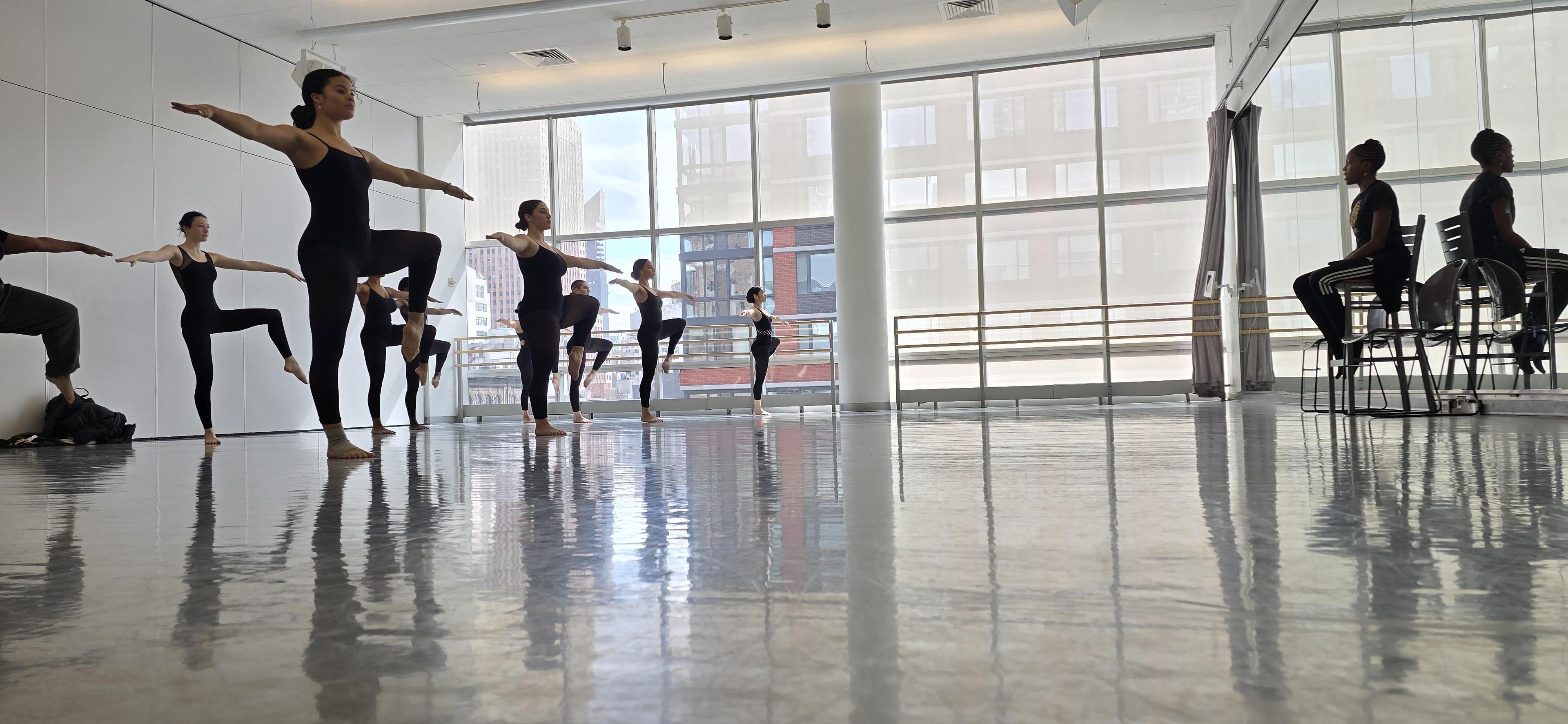 A group of dancers in black leotards are standing on one leg in two lines in a dance studio. Near the mirror is a teacher in all black sitting on a chair.