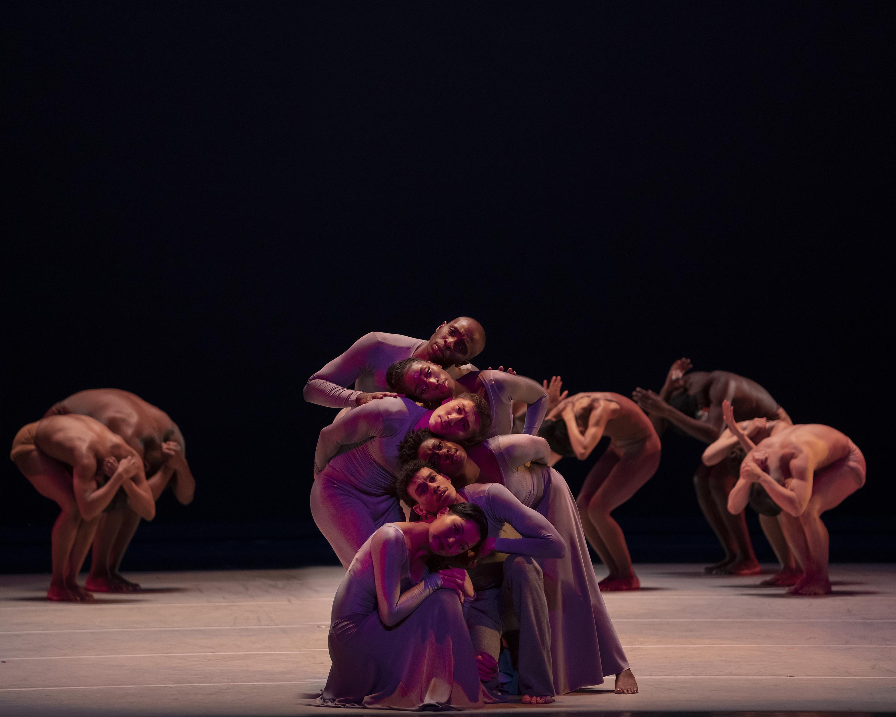 Dancers on stage are posed with their knees bent and heads on one another. They form three lines. They are all in drapey brown dresses.