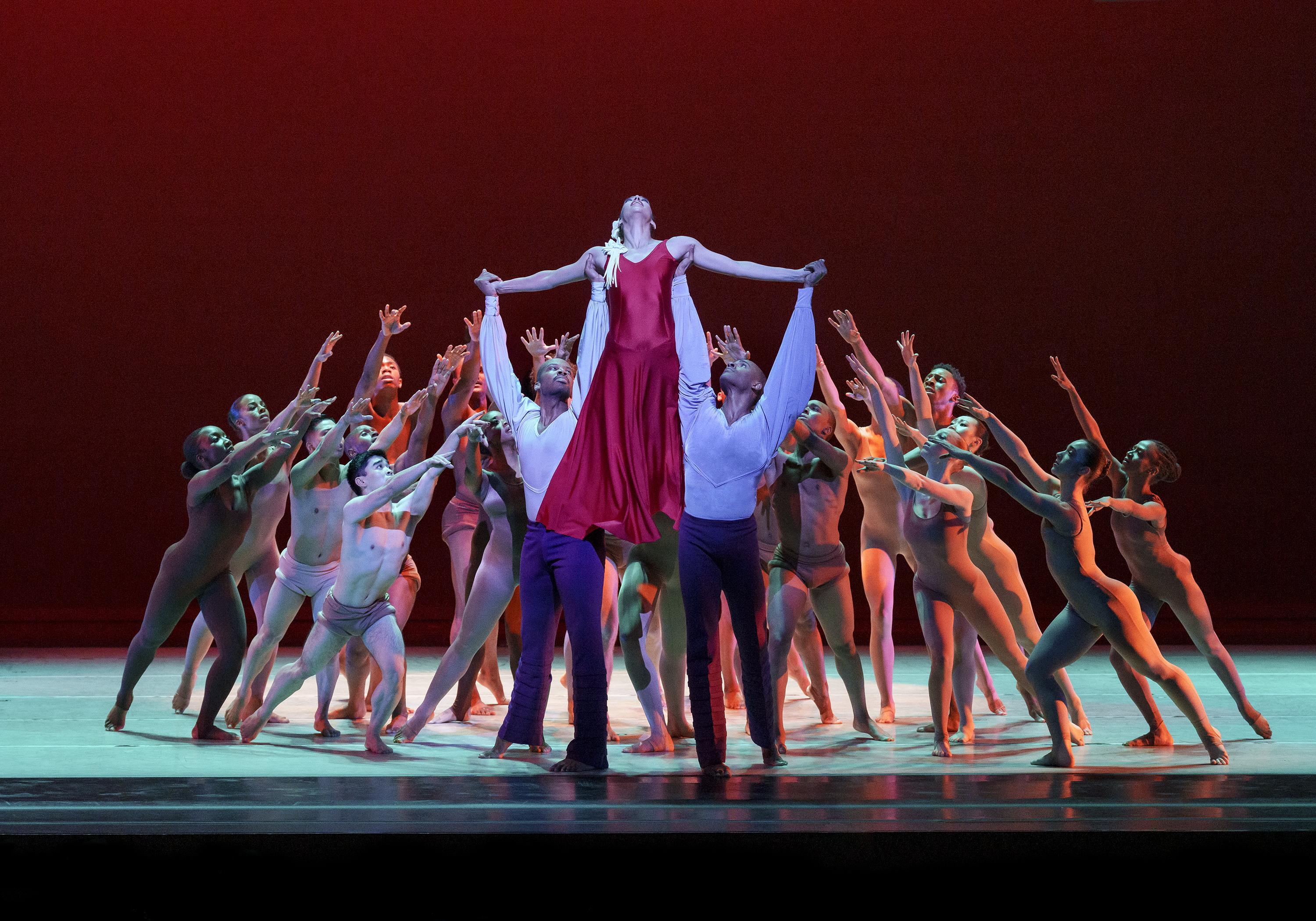 A group of dancers are huddled on stage forming a circle with their arms raised towards two men holding up one woman in a cross-like position. The woman is in red and the men are in white button downs and black pants.