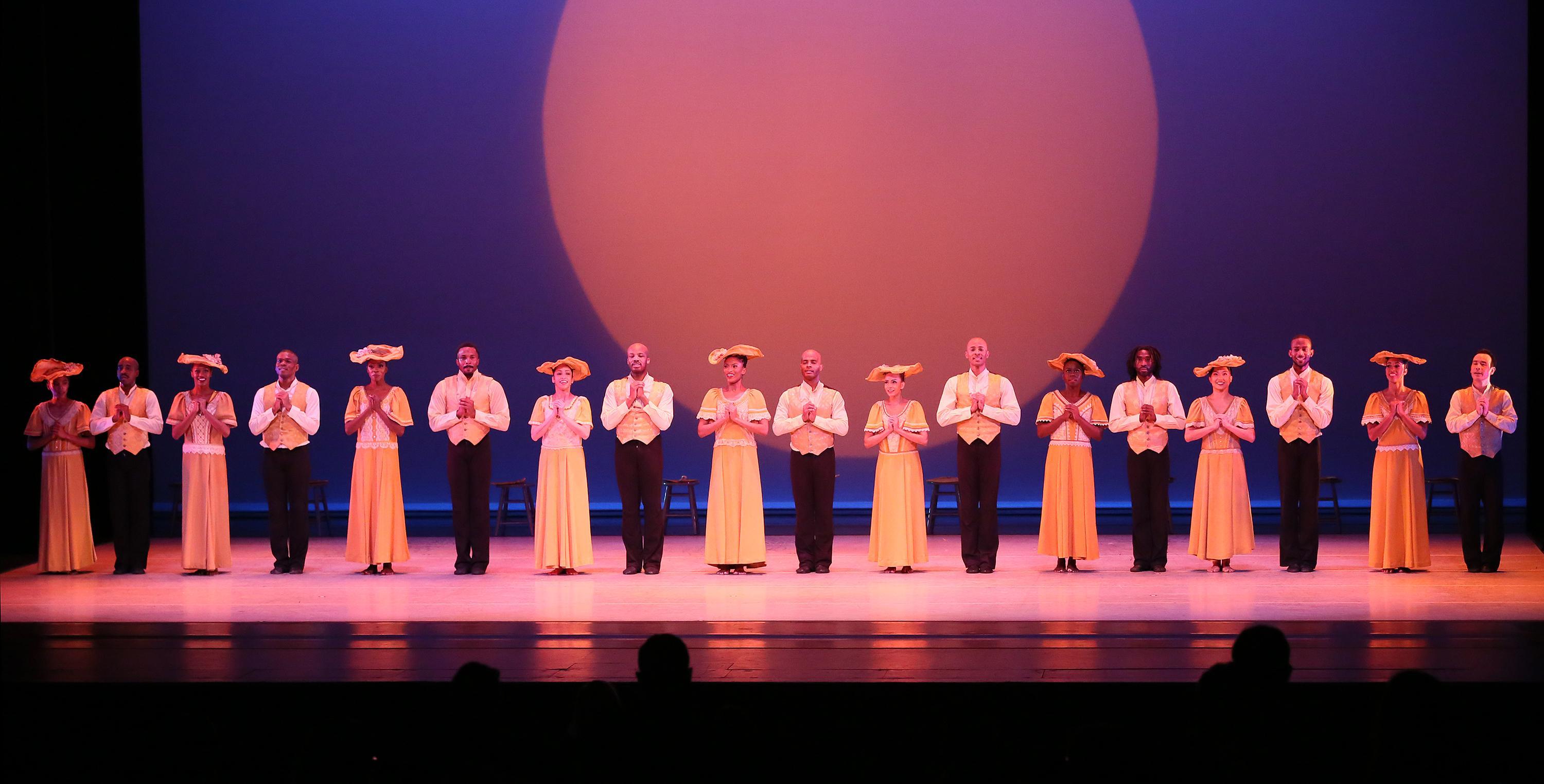 A group of dancers on stage, alternating men and women. The men are in black pants and white button down shirts. The women are in yellow dresses with hats.