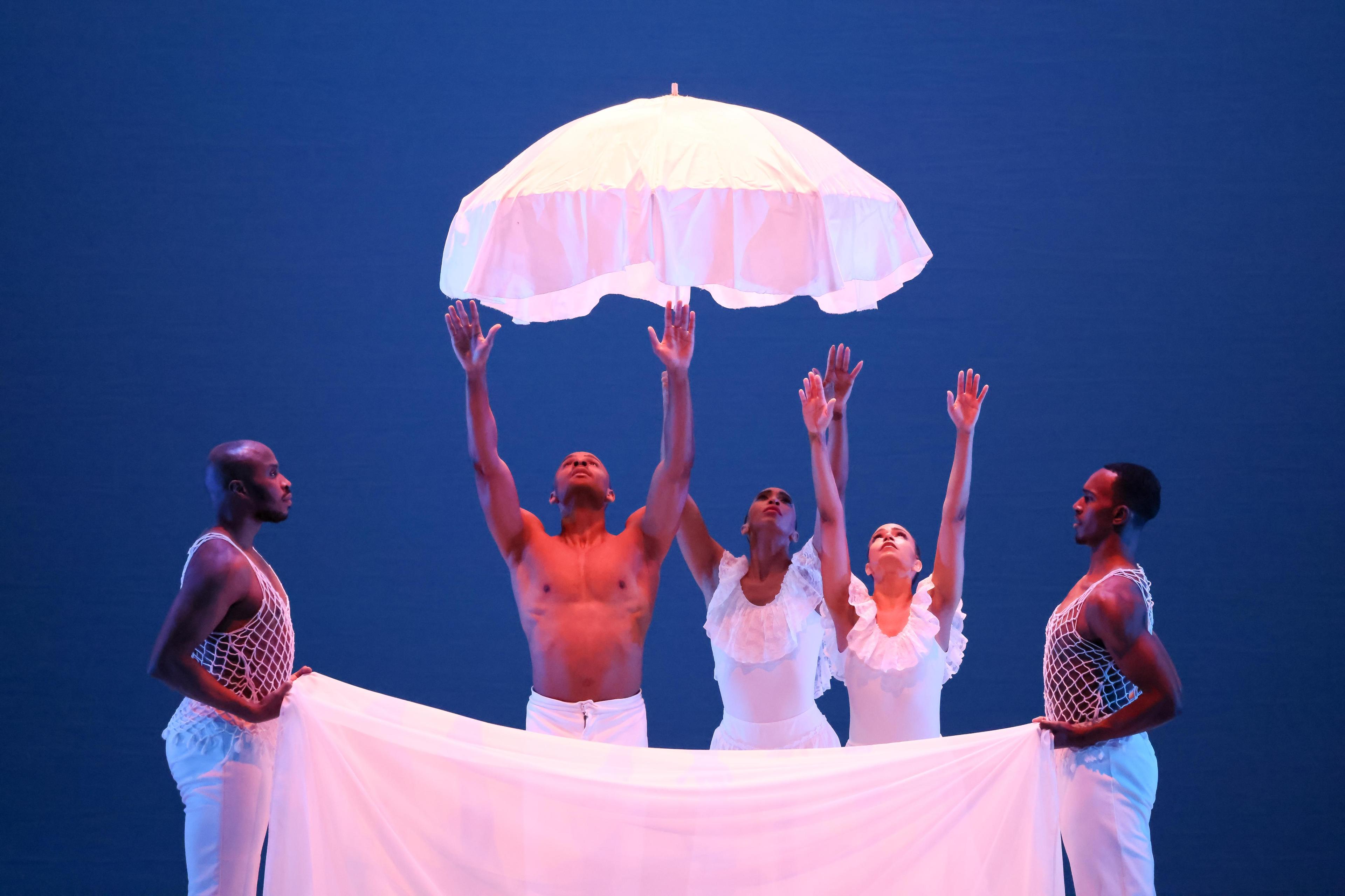 Two dancers are holding a sheet. Behind the sheet are 3 other dancers with their arms raised up to an umbrella.