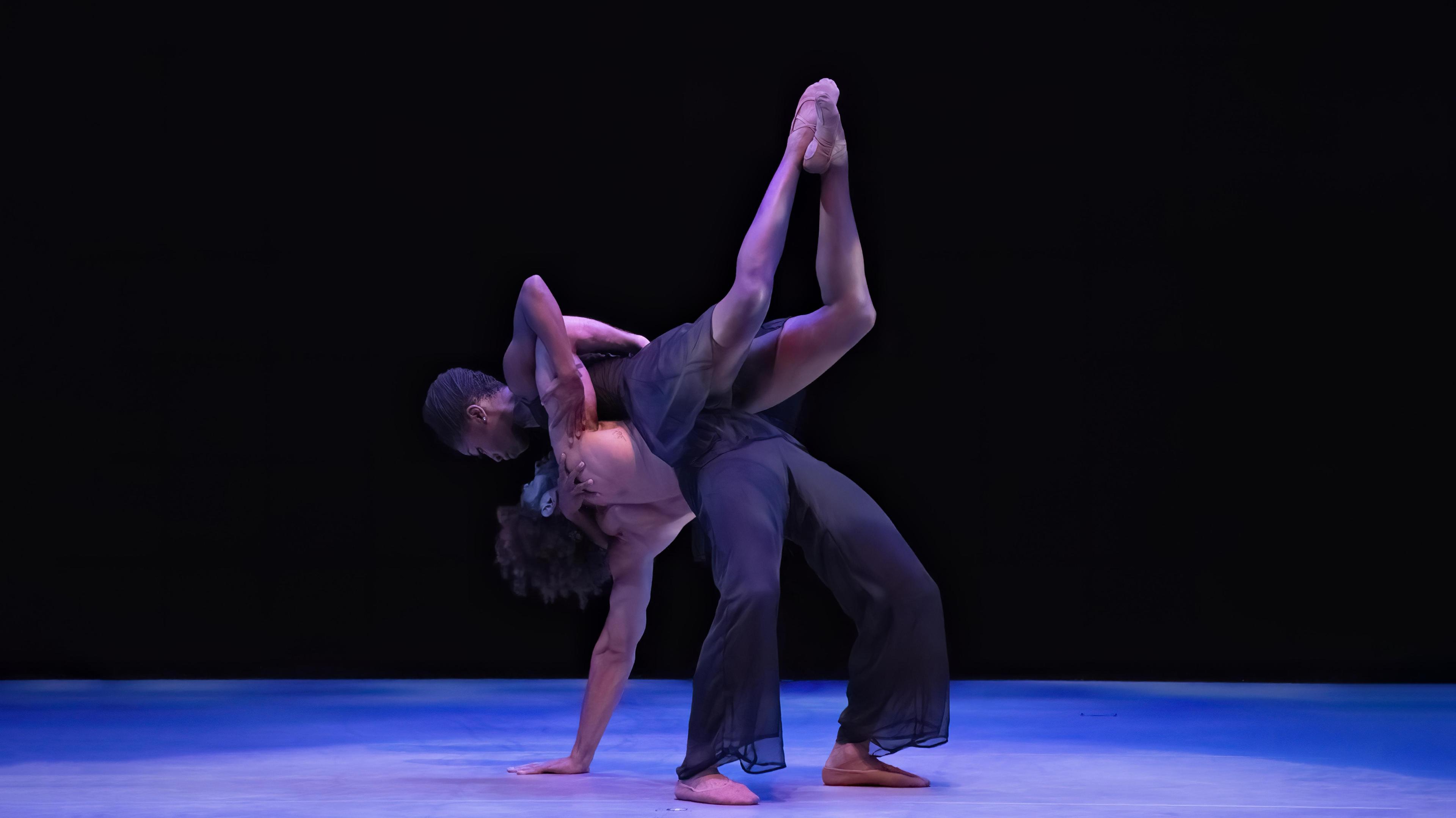 A man in a backbend with one hand on the ground and a women rests on his chest with her legs bent up in the air.