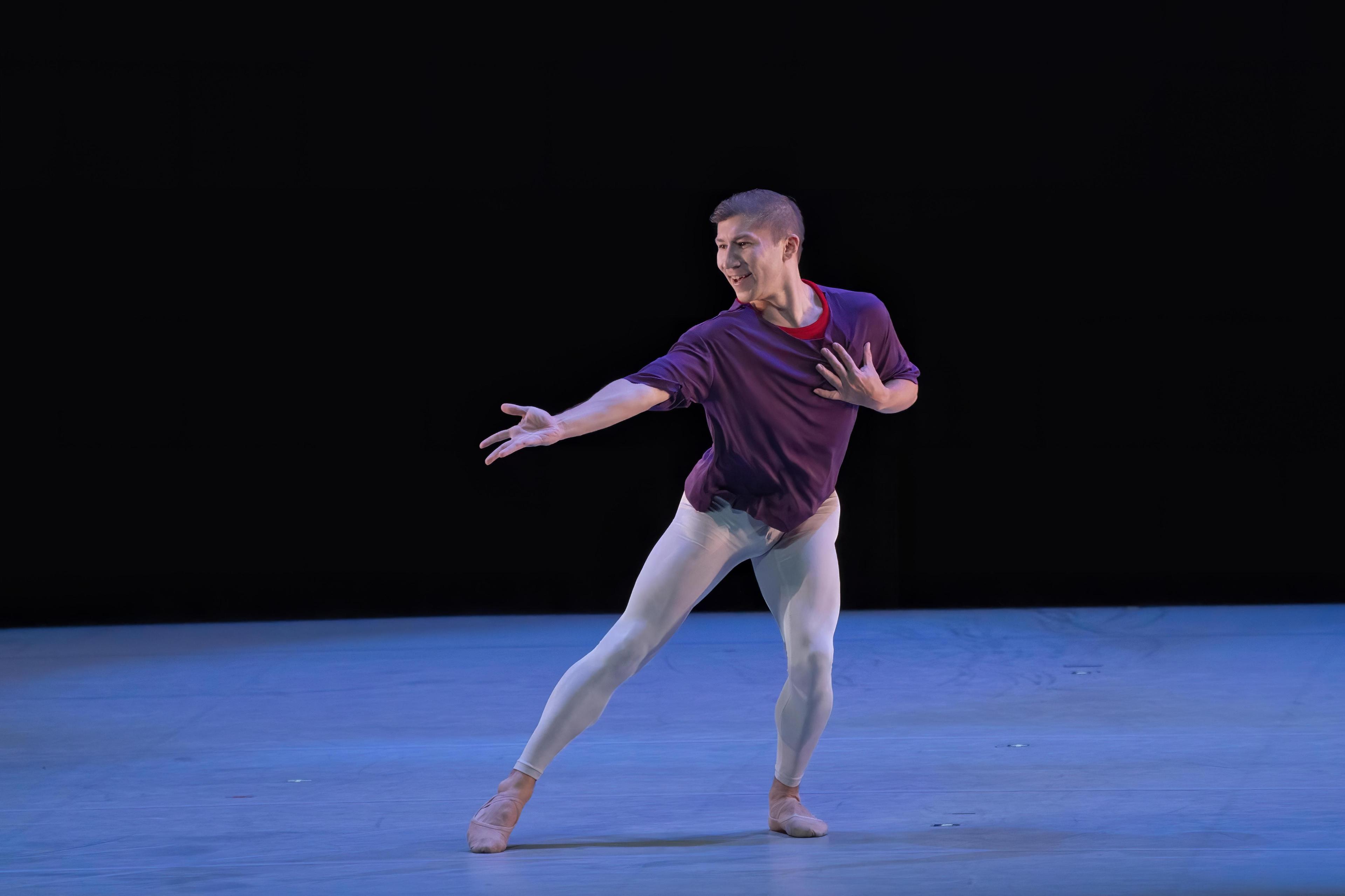 A man in a purple shirt and blue tights stans with one leg and one arm outreached.