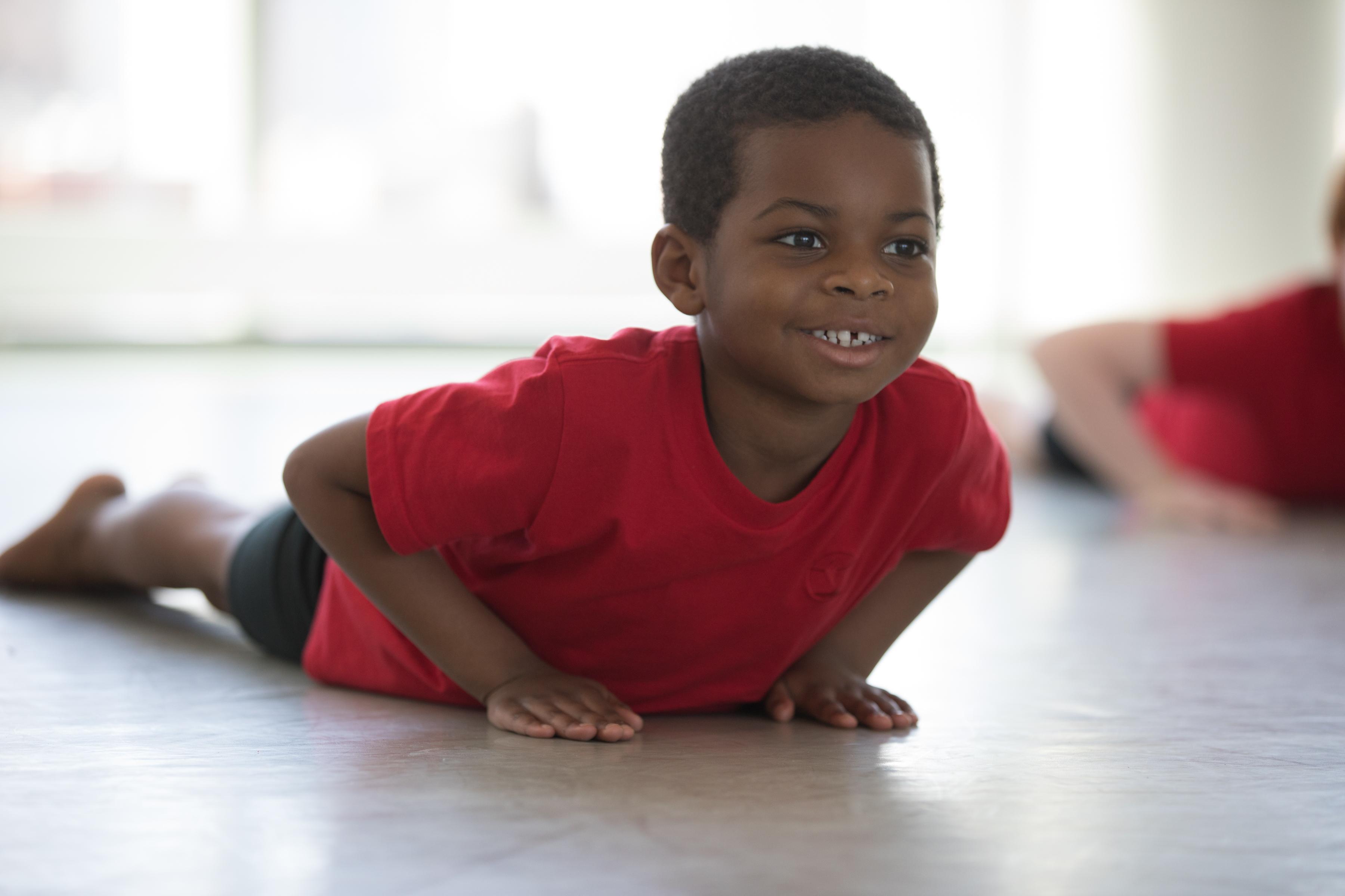 A boy in a red t-shirt laying on the ground on his stomach, looking up and smiling