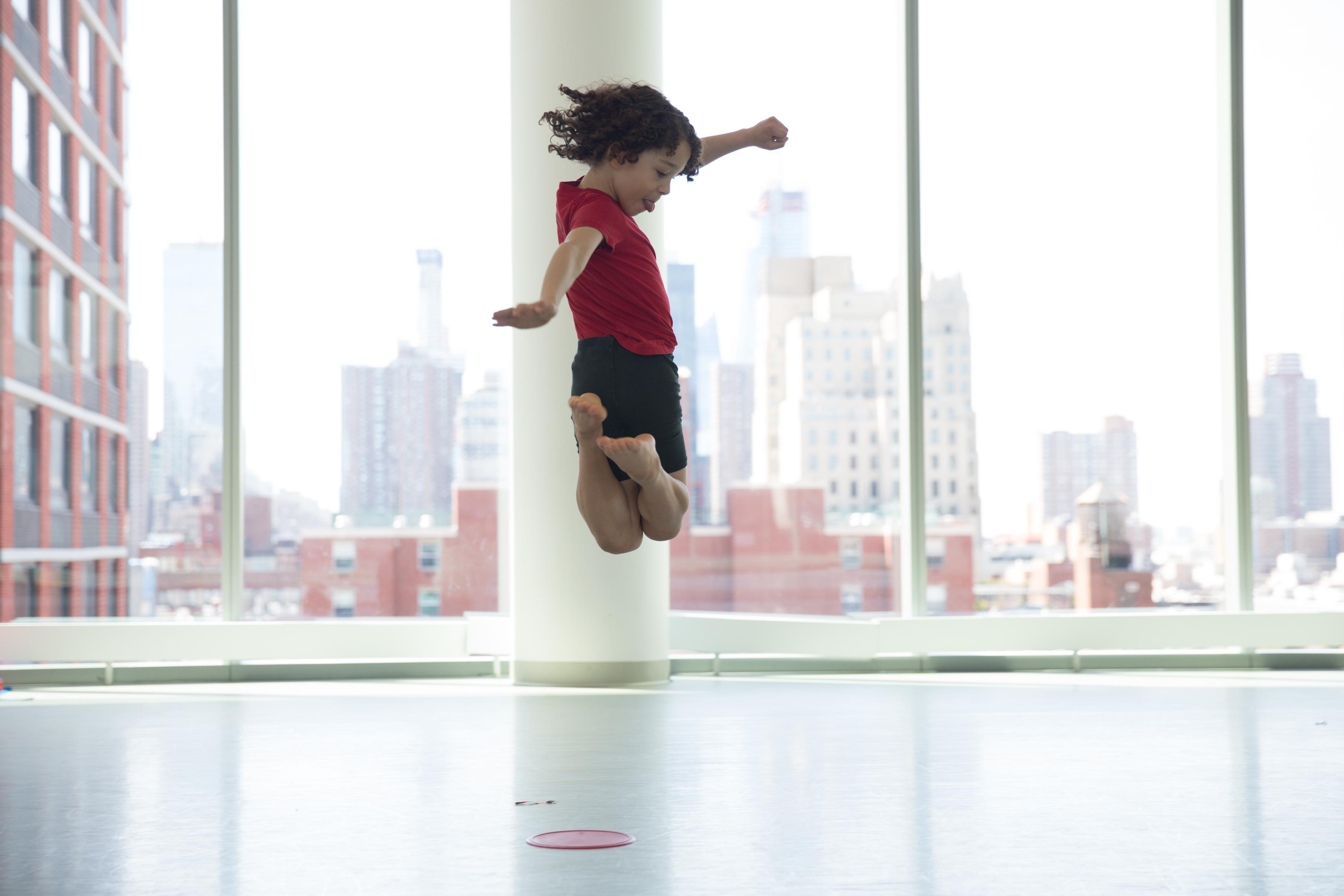 boy in red shirt and black shorts jumping in the hair with his feet tucked underneath him.