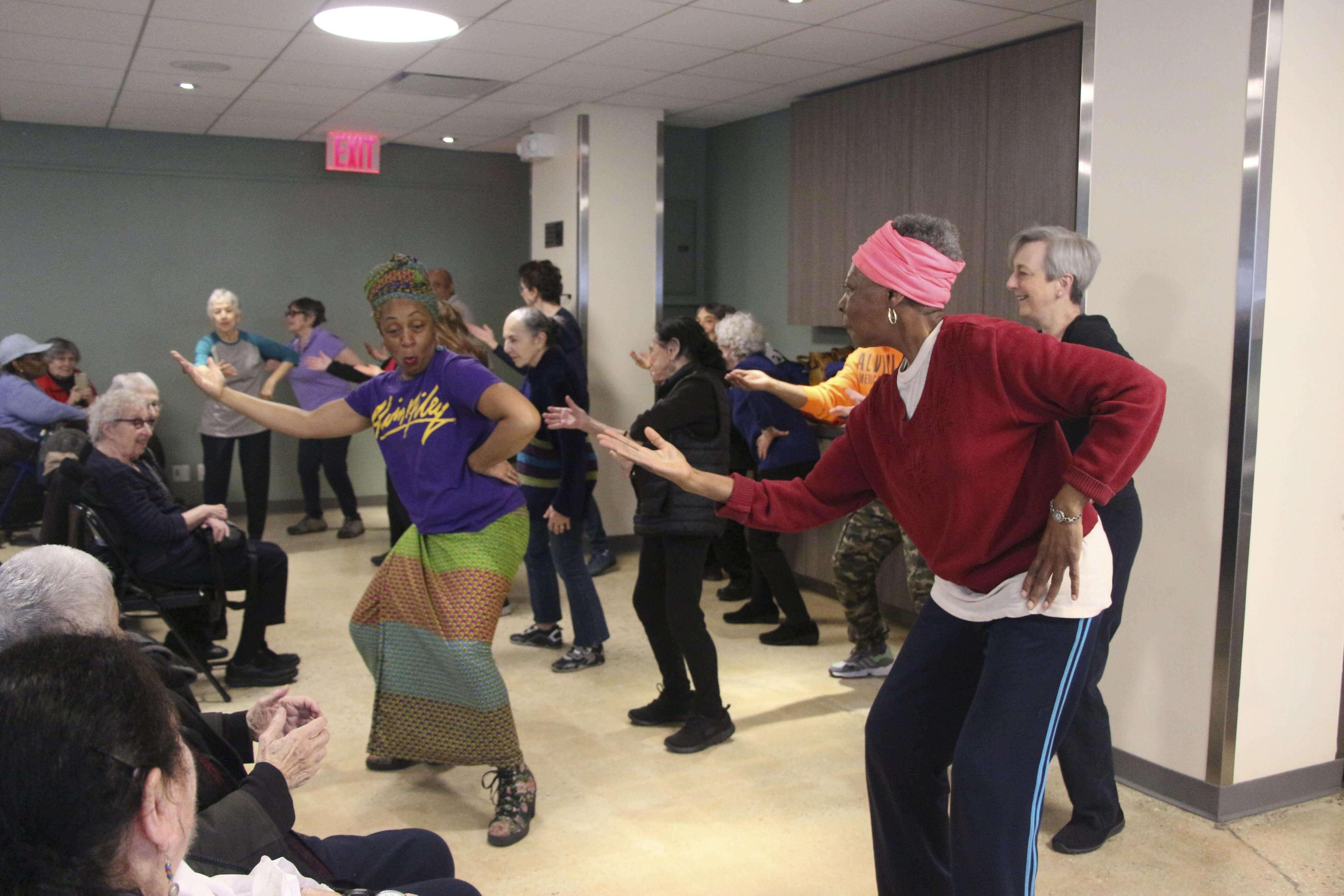  A group of seniors participating in a dance class at Penn South, led by an instructor in a purple Ailey T-shirt and colorful skirt. The seniors are smiling and actively engaged in the dance movements. The room is bright with overhead lighting, and some attendees are seated, watching the demonstration.