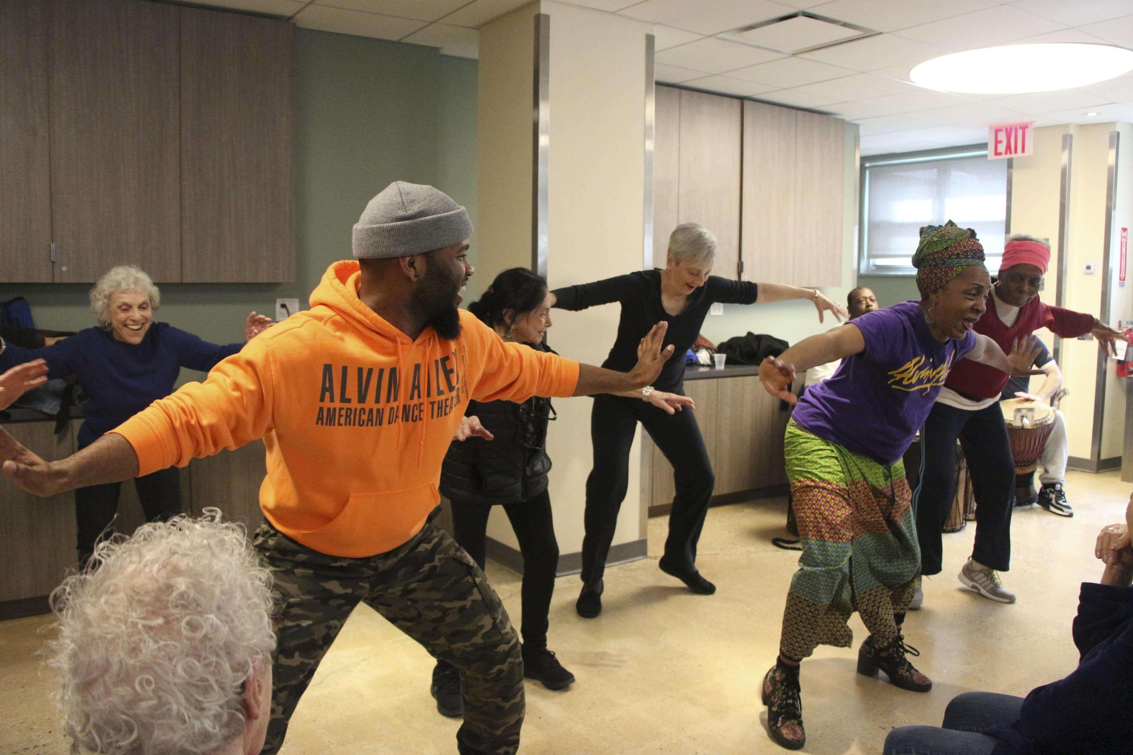 Seniors participating in a lively dance class at Penn South, led by an instructor in a purple Ailey T-shirt and colorful skirt, and another instructor in an orange Alvin Ailey hoodie and camo pants. The participants are smiling and engaged in dance movements, with some attendees watching from their seats. The room is brightly lit with overhead lighting.