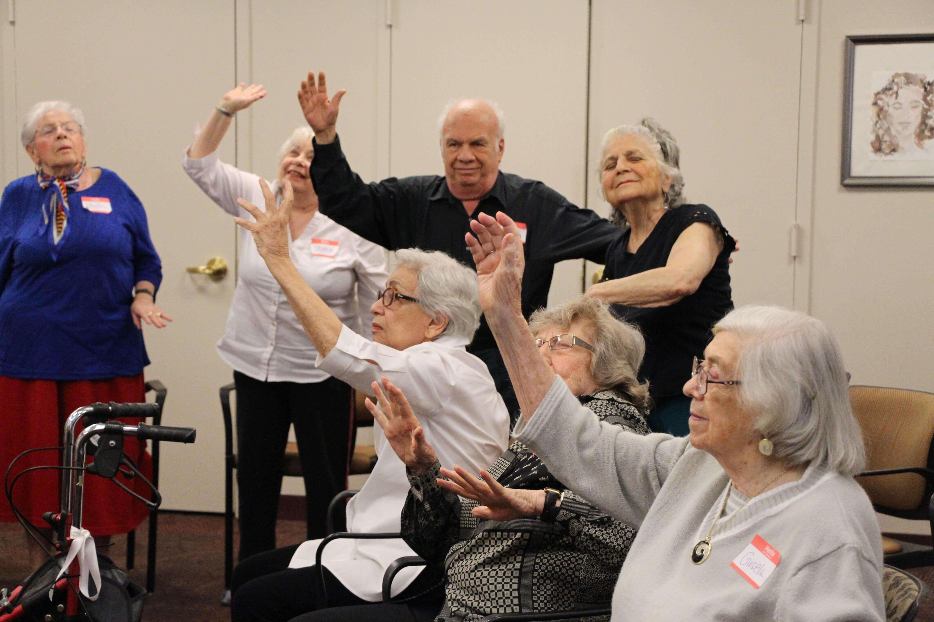 Seniors participating in a seated dance class at Carnegie East, raising their arms and smiling. Some participants are standing while others remain seated, all engaging in the movements. The room has a warm and welcoming atmosphere.