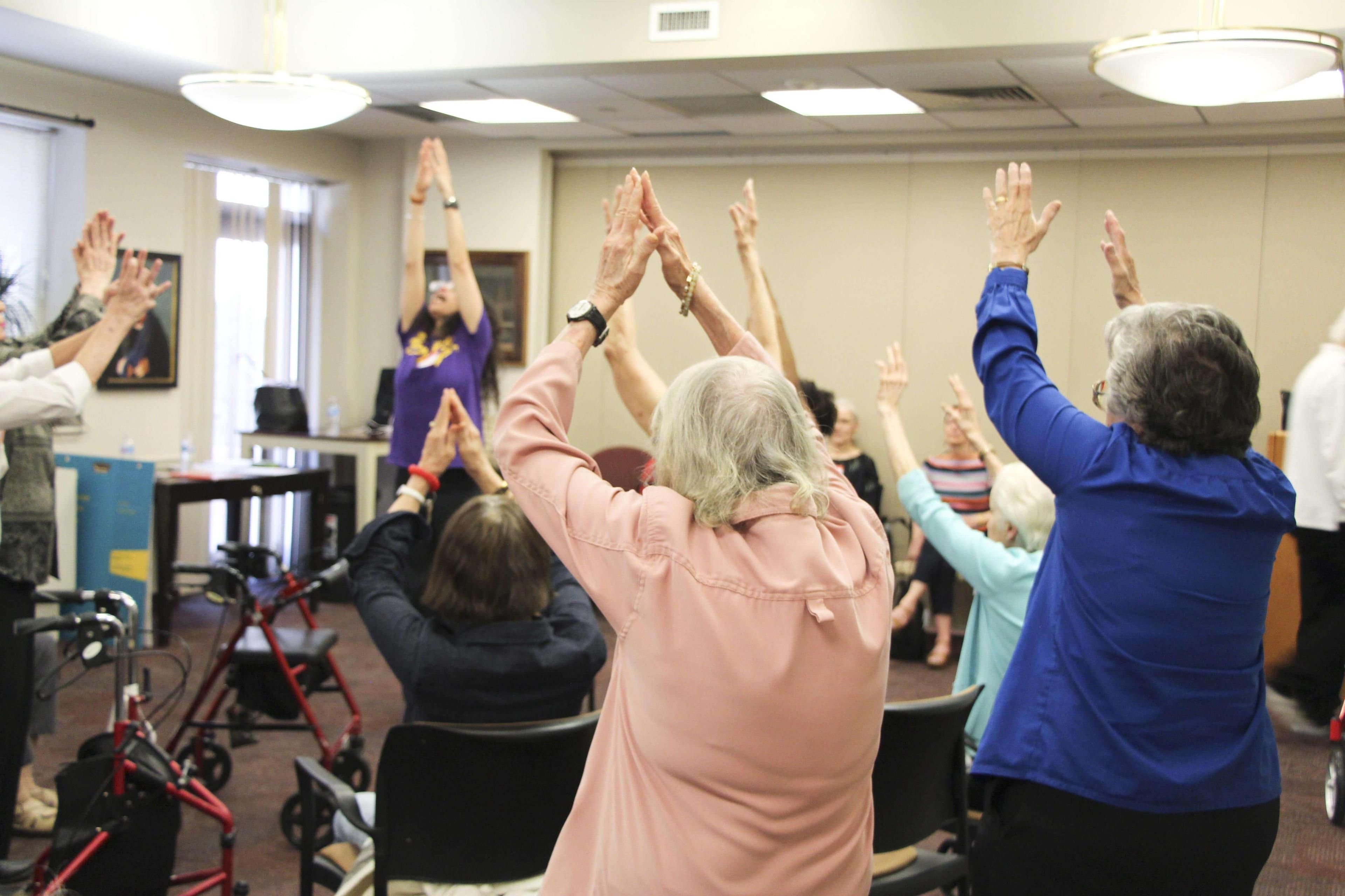  Seniors in a seated dance class at Carnegie East, raising their arms in unison. An instructor in a purple Ailey T-shirt leads the class from the front. The room is well-lit with large windows, and several participants use walkers or are seated in chairs.