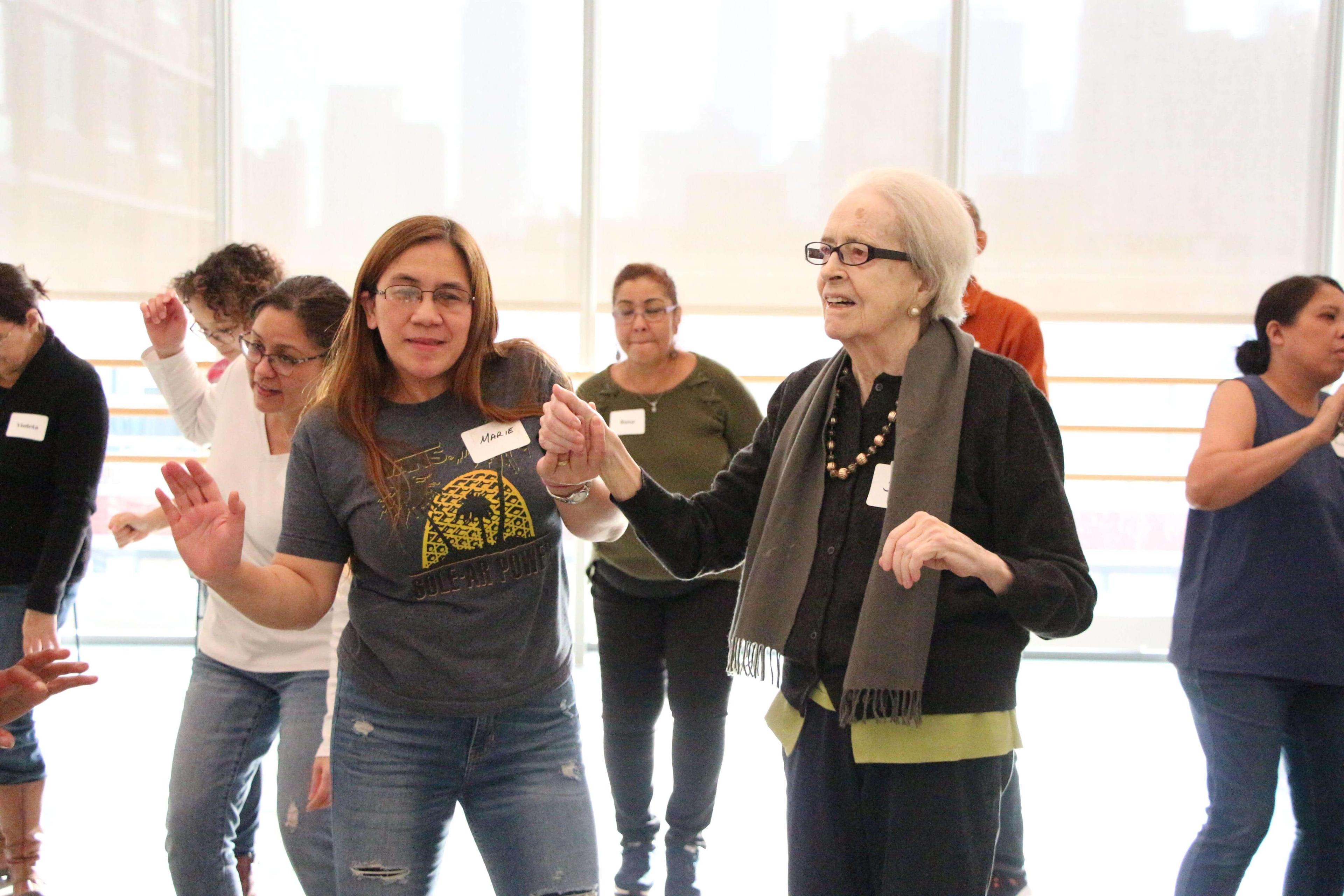 Group of people dancing in a bright room. An elderly woman with white hair and glasses, wearing a black outfit and scarf, is holding hands with a younger woman in a gray shirt and jeans. The young woman's name tag reads Maria. Other participants, including a woman with a name tag reading Guadalupe, are visible in the background, all smiling and dancing.