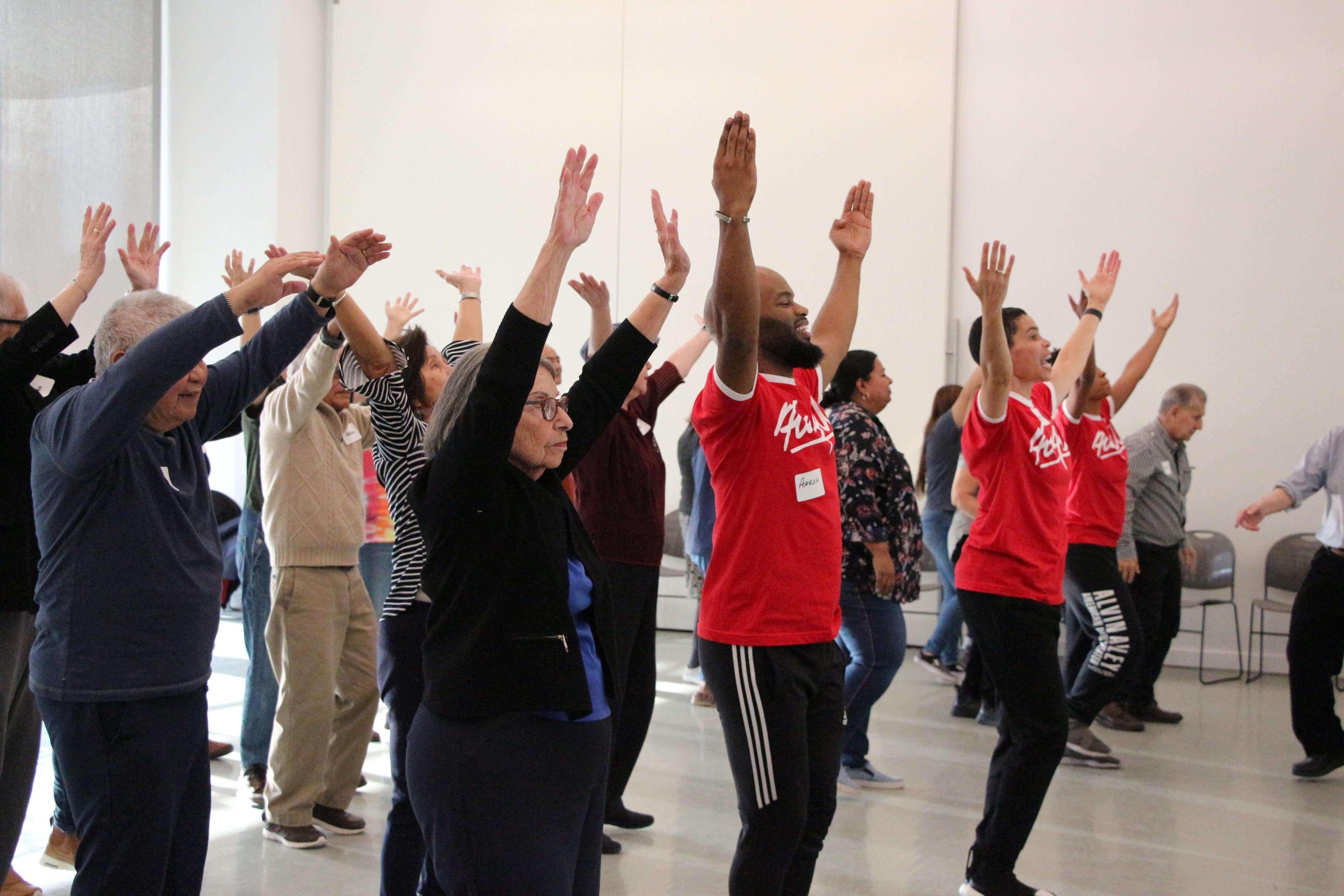 Group of people dancing with arms raised in a bright room. Participants include seniors and younger individuals, all following the movements of leaders in red shirts. The leaders' shirts say Flam Riley, and one leader's name tag reads Aaron. Everyone is engaged and enjoying the activity.