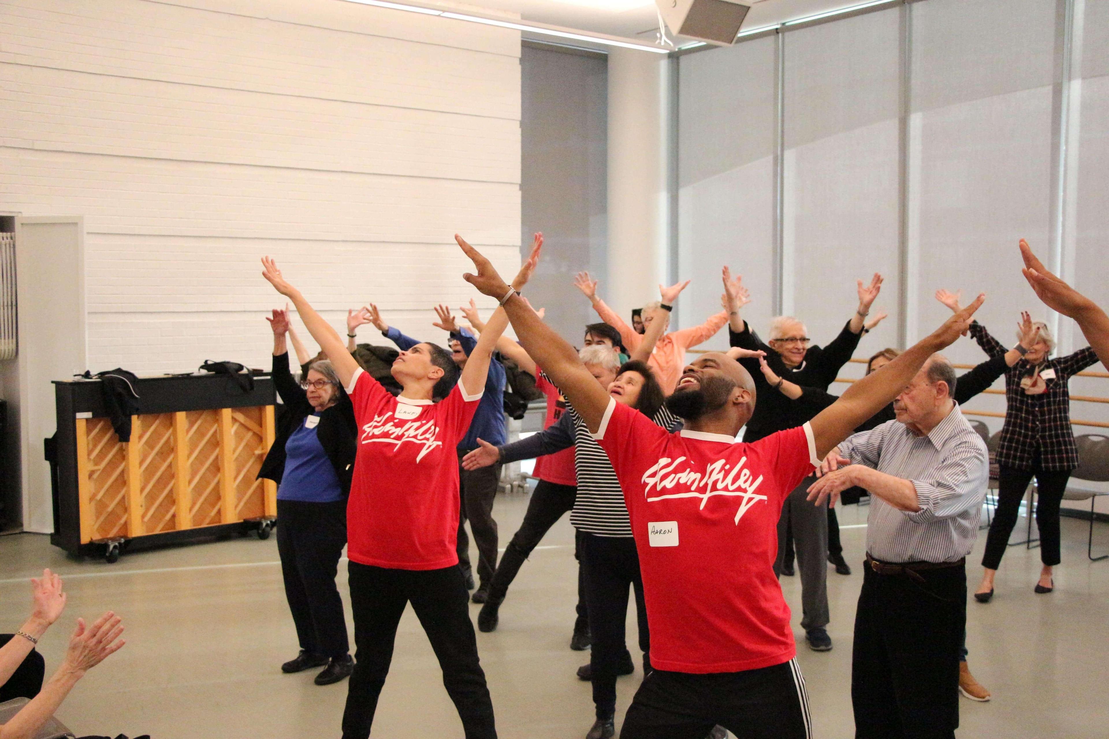  Group of people dancing with arms raised in a bright room. Two men in the foreground, wearing matching red shirts that say Flam Riley, lead the group. One man’s name tag reads Aaron. Participants of various ages and backgrounds follow their movements, smiling and enjoying the activity. A piano is visible in the background.