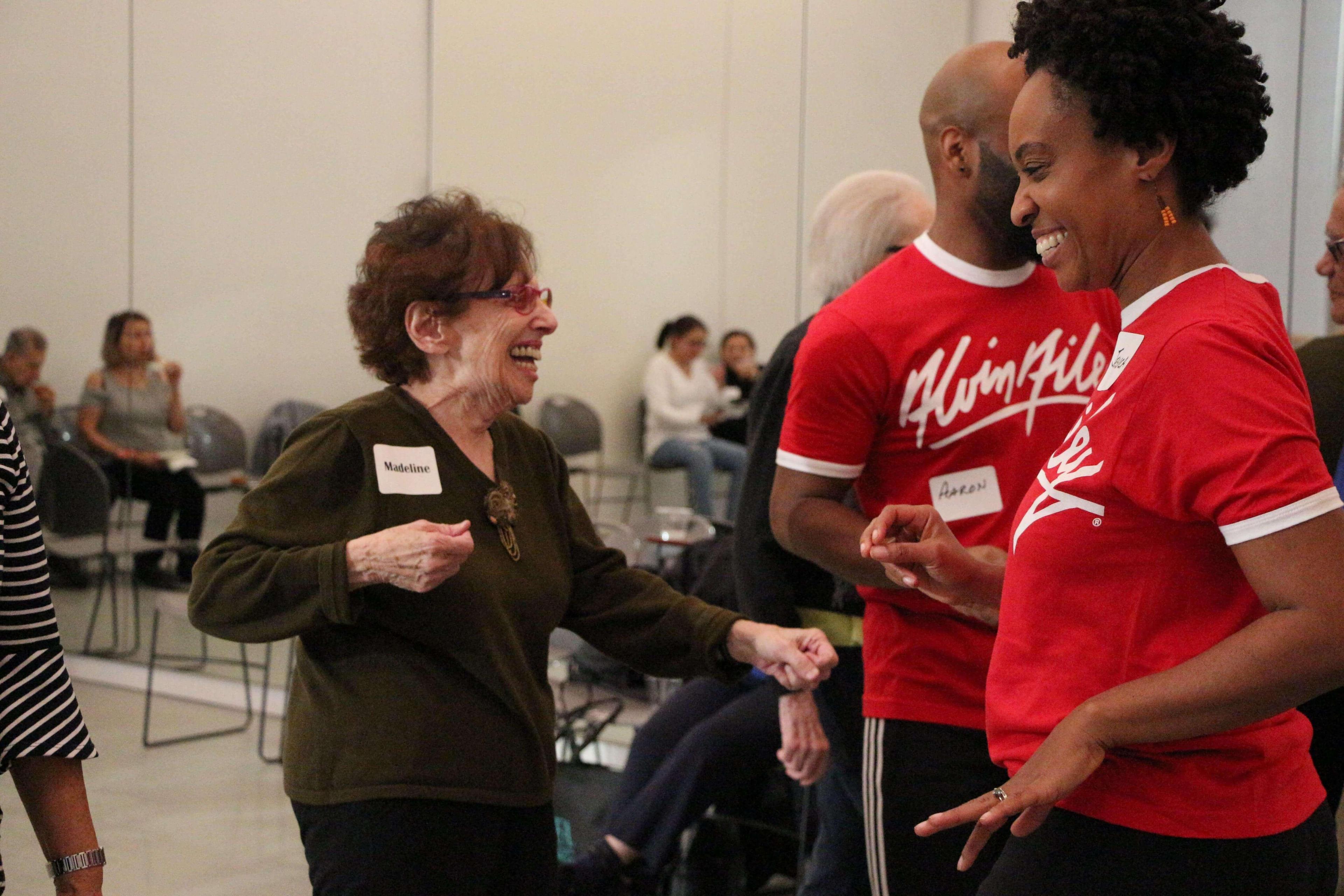 An elderly woman with a name tag reading Madeline is smiling and dancing with two leaders wearing red shirts that say Flam Riley. One leader's name tag reads Aaron. They are all laughing and enjoying the moment. Other participants are seated in the background.