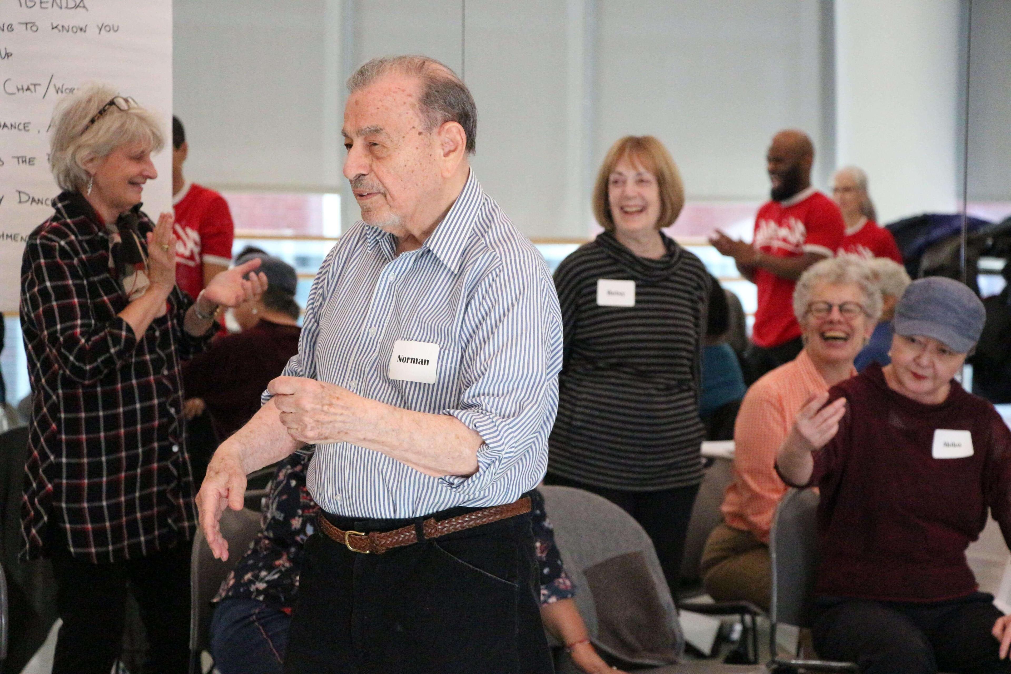 Seniors participating in an intergenerational dance party with the NYU Family Support Program. A man named Norman, identified by his name tag, dances in the foreground, while other participants, including women named Reba and Shirley, smile and clap in the background. The room is bright and filled with natural light.