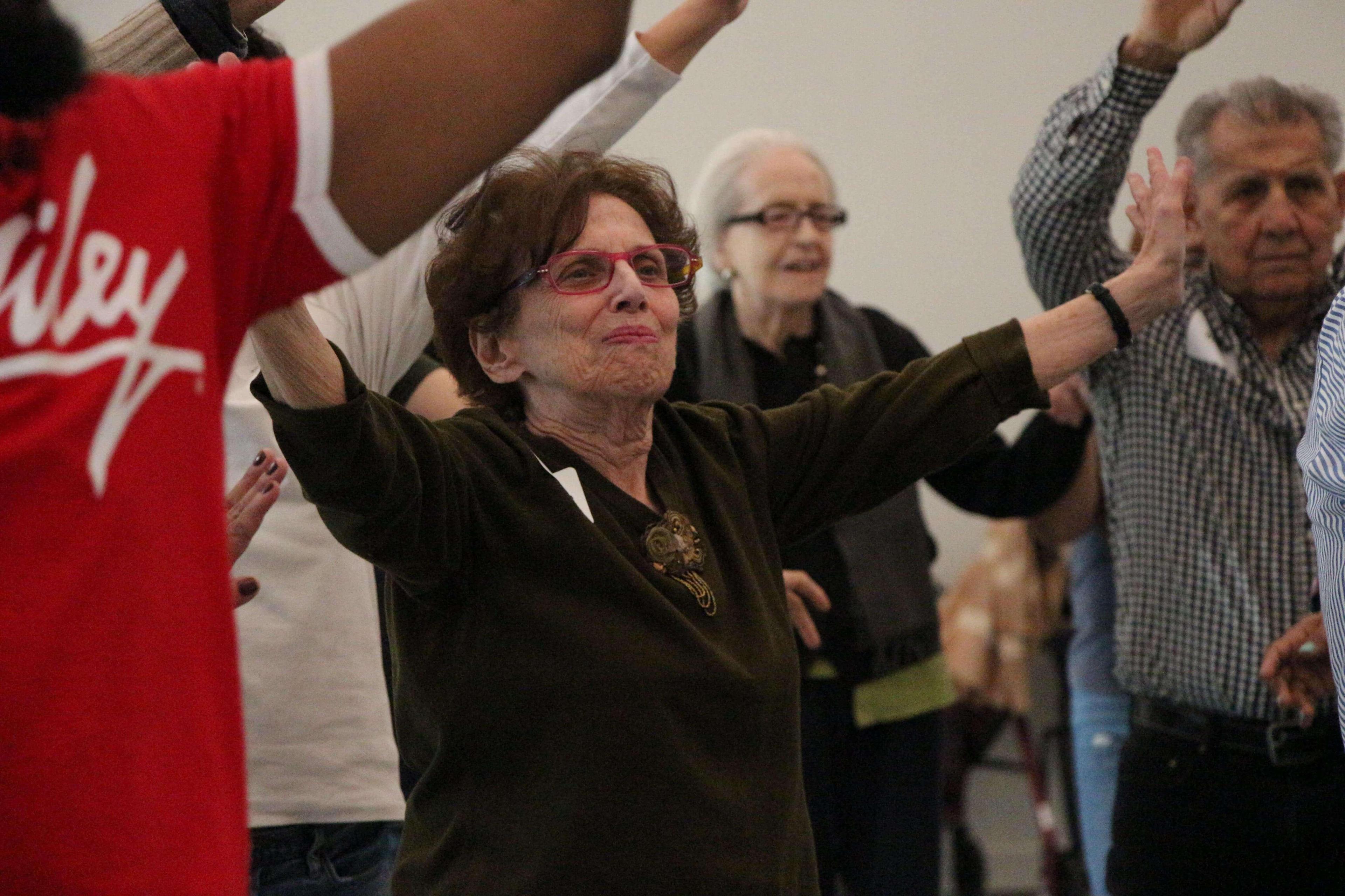  Seniors participating in an intergenerational dance party with the NYU Family Support Program. An elderly woman in the foreground, wearing glasses and a dark top, raises her arms with a focused expression. Other participants, including a man and woman in the background, are also engaged in the dance. The room is lively and filled with energy.