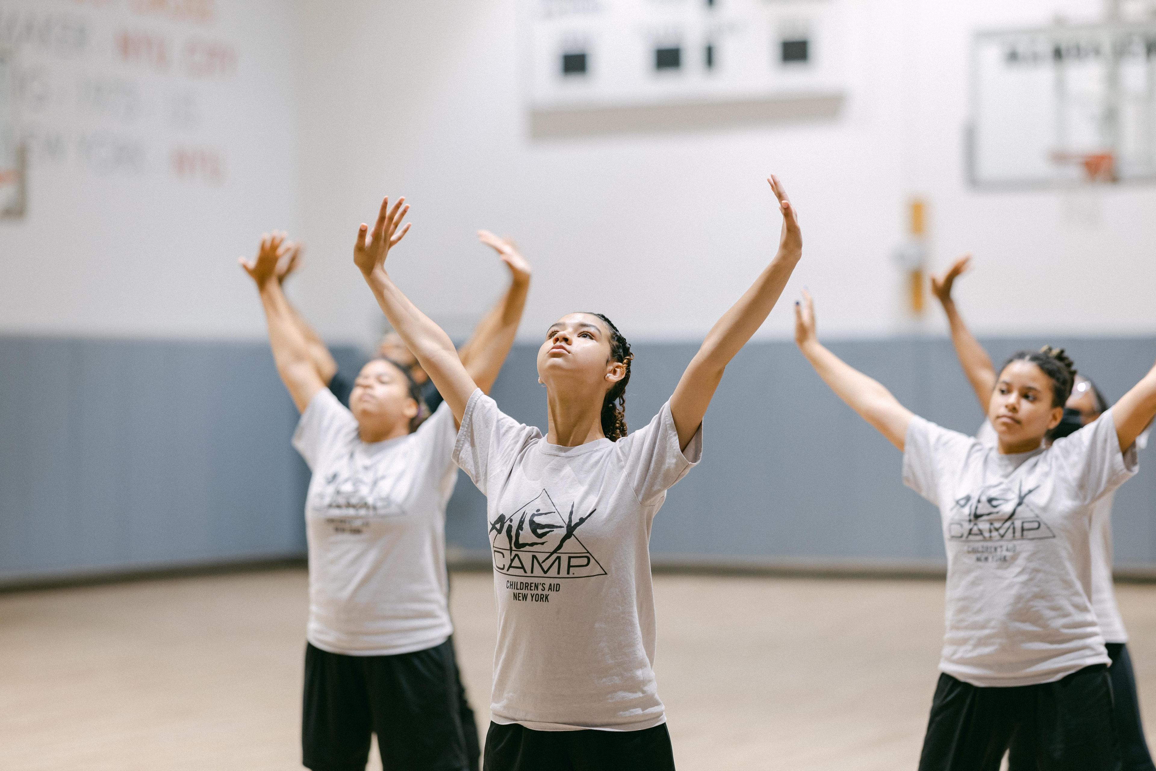 3 students in AileyCamp tshirts with arms upward and turned outward