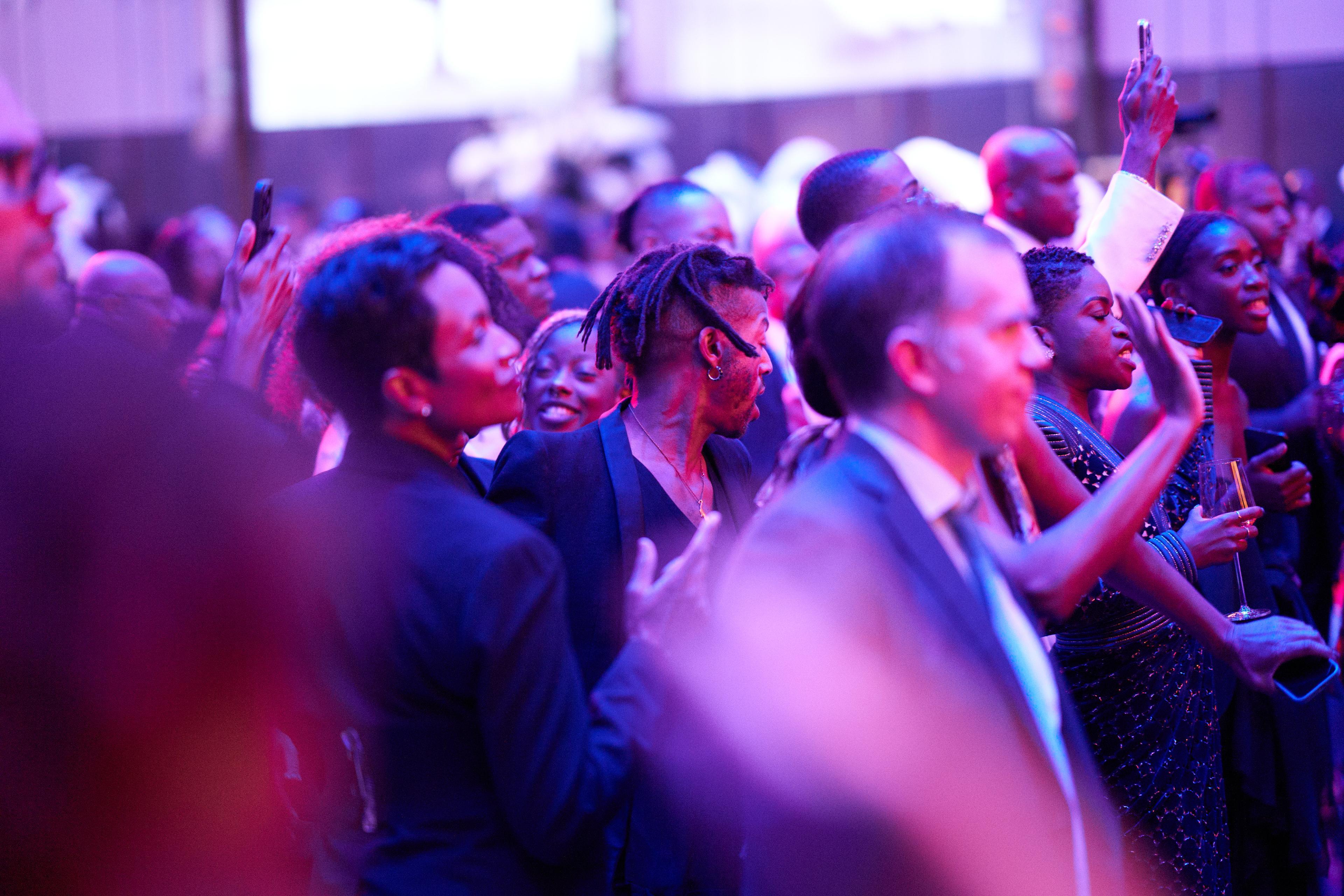 A room full of people dancing in a ballroom. There is a pink tint to the picture due to lights from the stage.