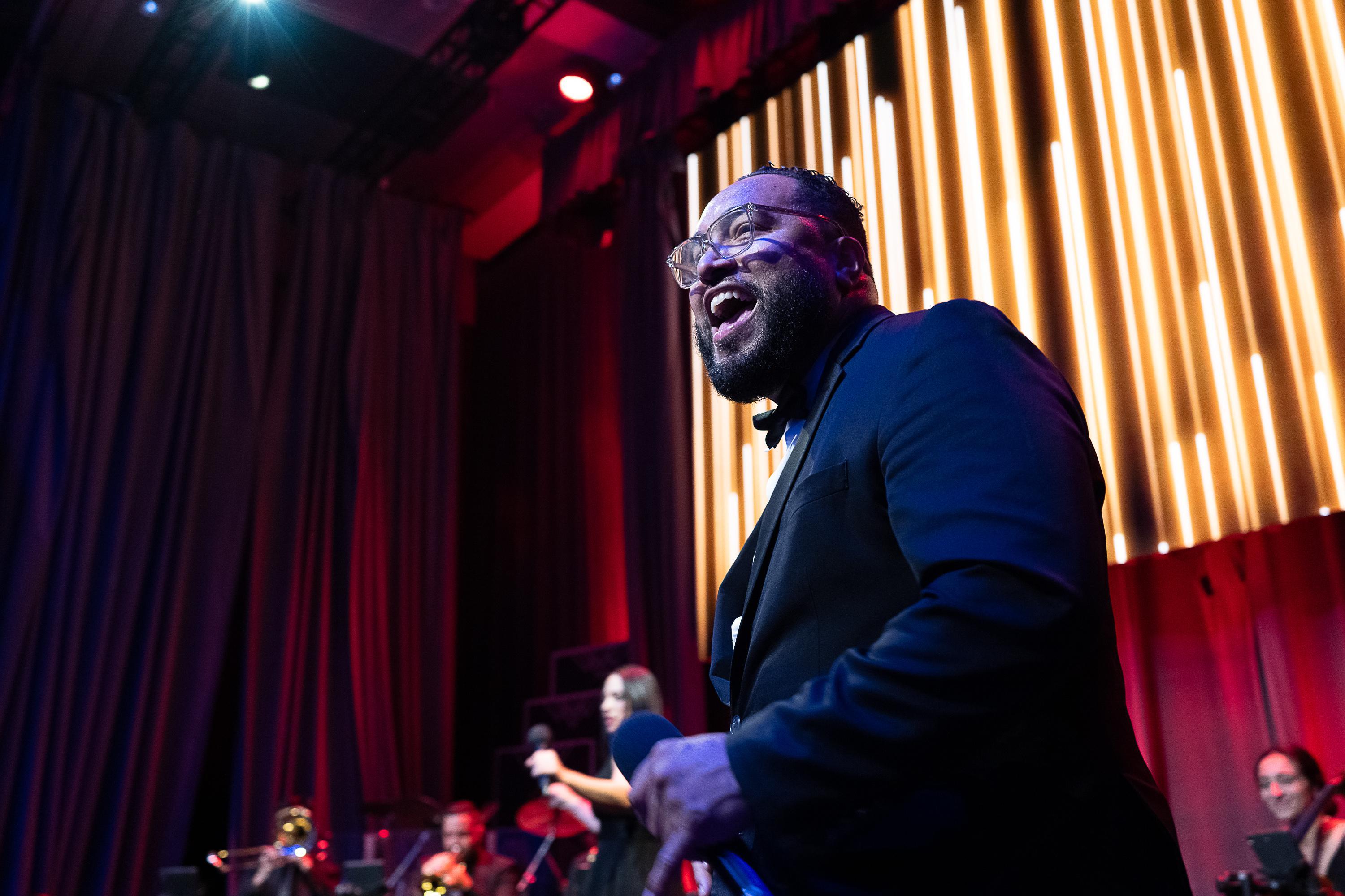 A man on stage in a black suit. He is the lead singer of a band and he is smiling at the crowd.