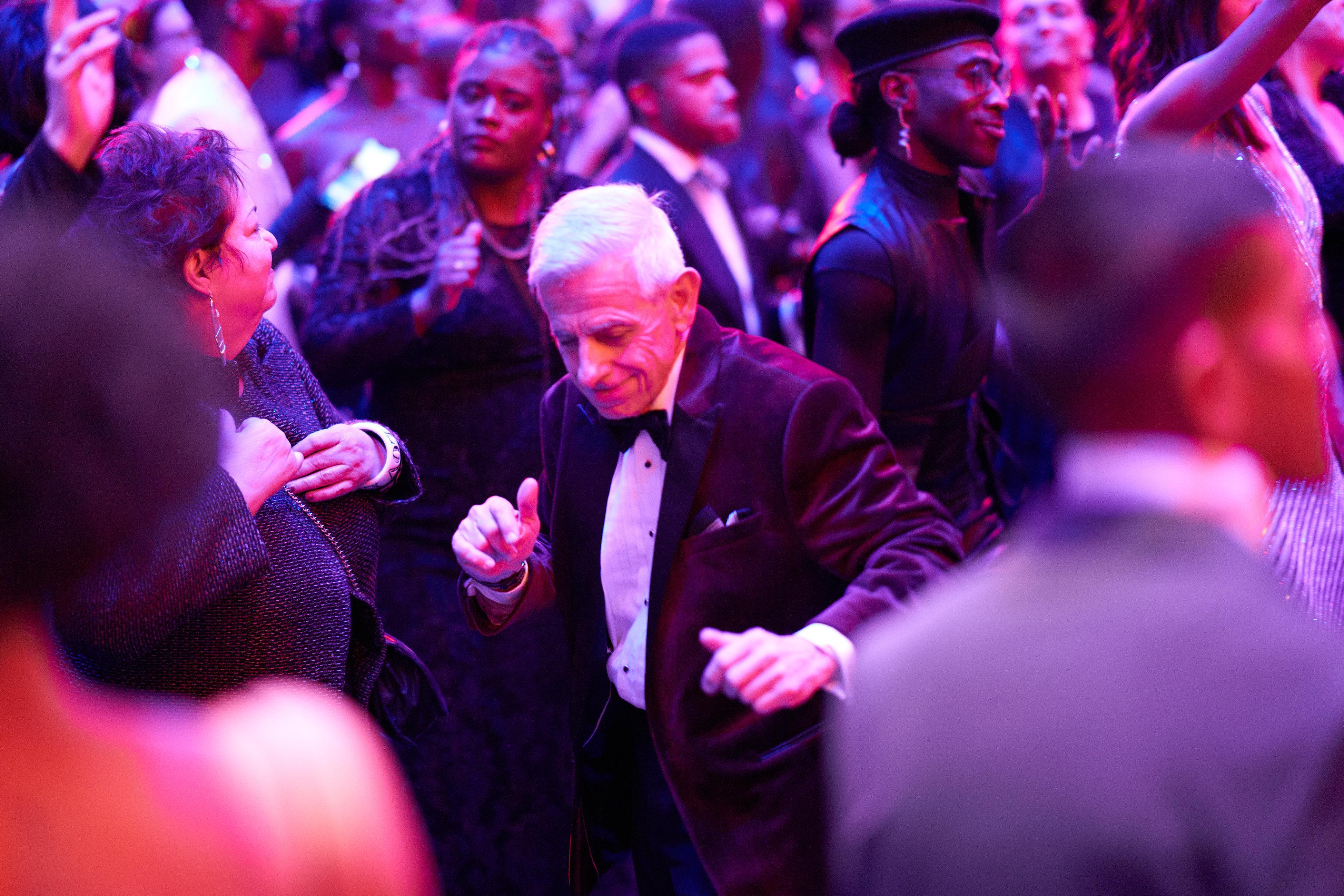 A group of people dancing in a ballroom. There is a man with grey hair and a tuxedo in the middle smiling and dancing.