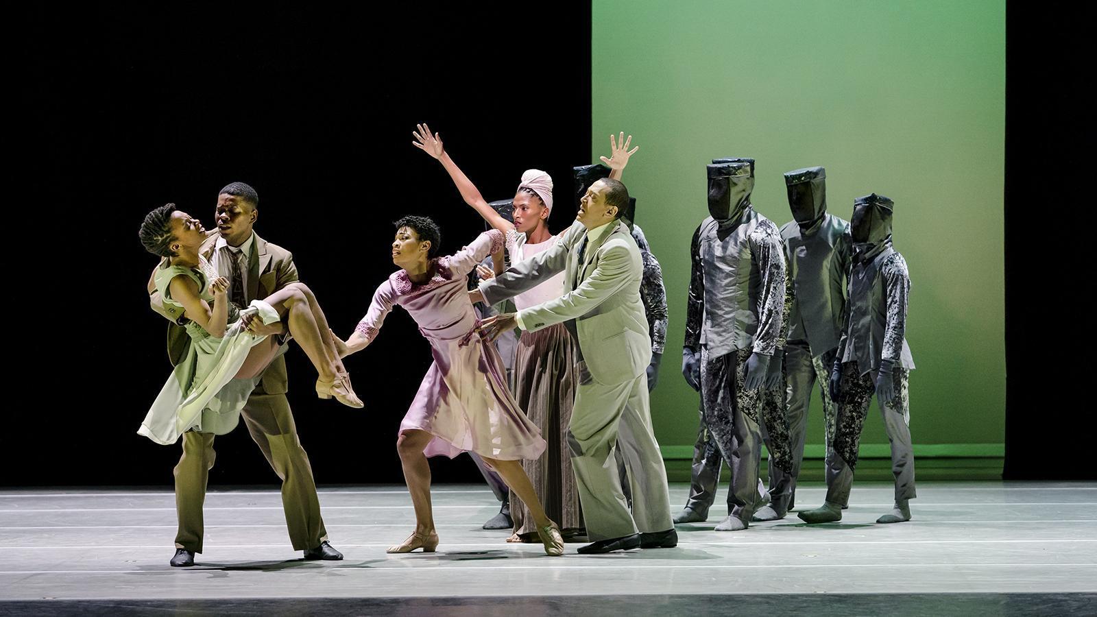 A group of dancers on stage performing a contemporary dance piece. One dancer is being carried by a male dancer, while another dancer in a pink dress extends her arms towards them. Three dancers in gray, futuristic costumes with covered faces stand in the background. The backdrop is lit with a gradient of green.