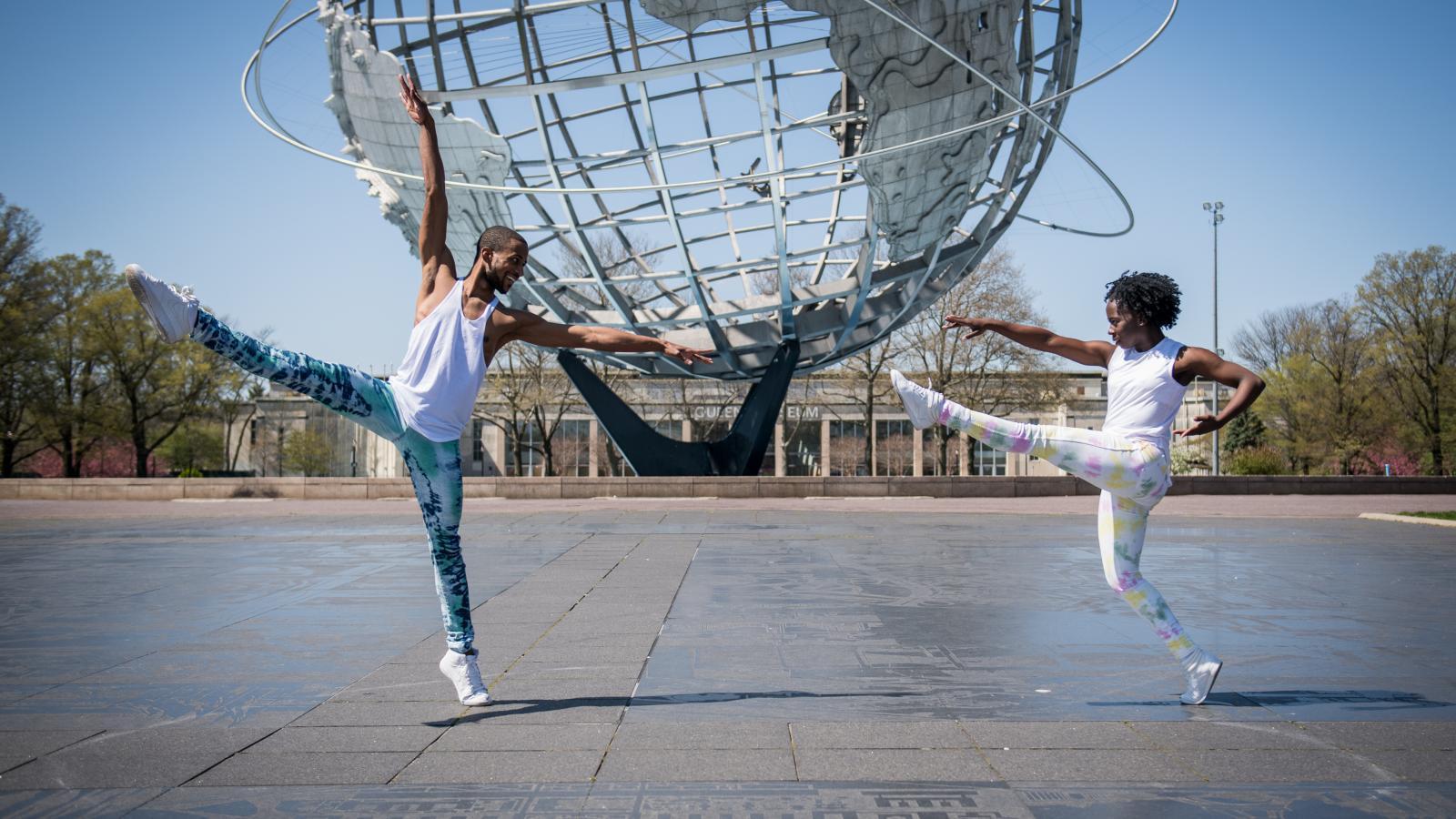 Two dancers, one male and one female, perform in front of a large metal globe sculpture. Both are wearing white tank tops and colorful leggings. The male dancer is balancing on one leg with the other leg and arm extended, while the female dancer mirrors his pose with a playful expression. Trees and a clear blue sky are visible in the background.