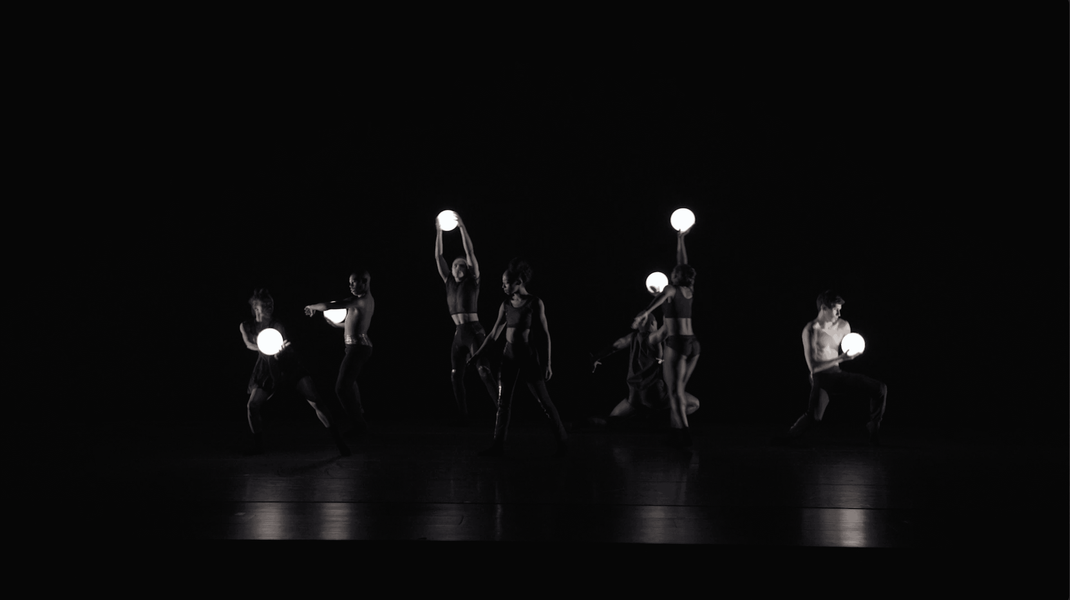  A group of dancers performs on a dark stage, each holding a glowing white orb. They are positioned in various dynamic poses, with some standing and others kneeling or stretching, creating a visually striking composition against the dark background.