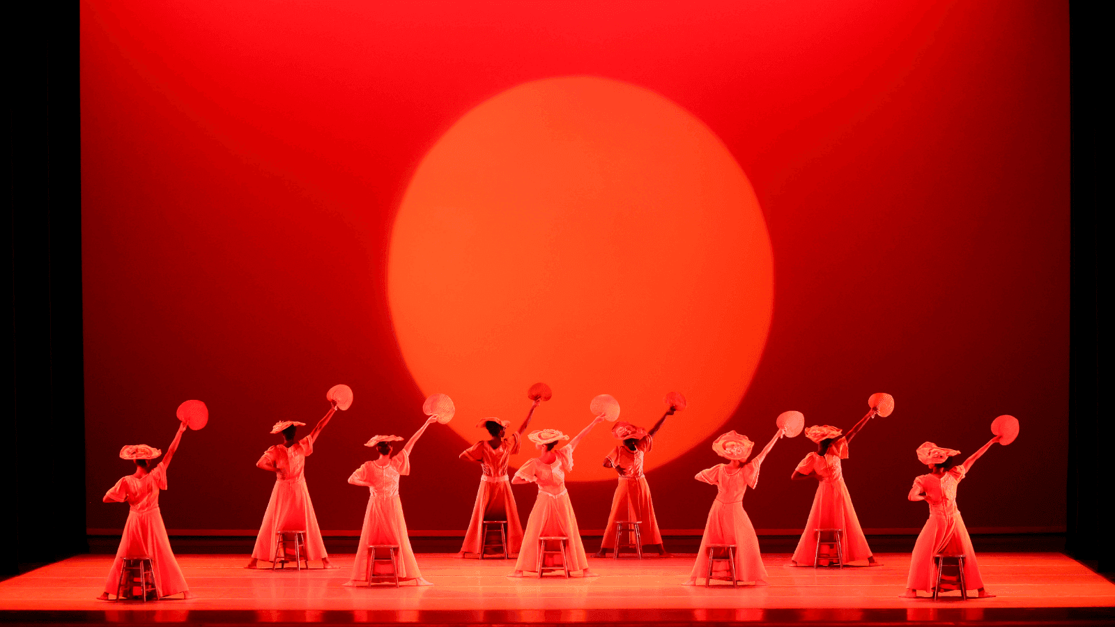  A group of dancers perform on stage in front of a large red sun backdrop. The dancers are dressed in red costumes, some holding fans, and are positioned behind stools with one arm raised. The stage is illuminated with intense red lighting.