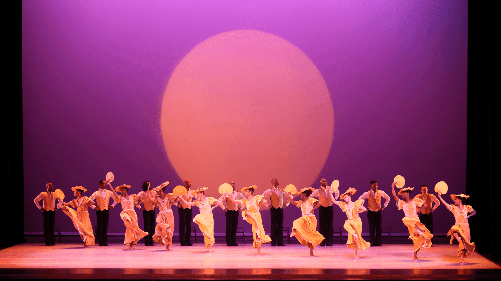 A group of dancers perform on stage in front of a large orange sun backdrop. The dancers are dressed in yellow and white costumes, some holding fans, and are mid-dance with expressive poses. The stage is bathed in warm orange and purple lighting.